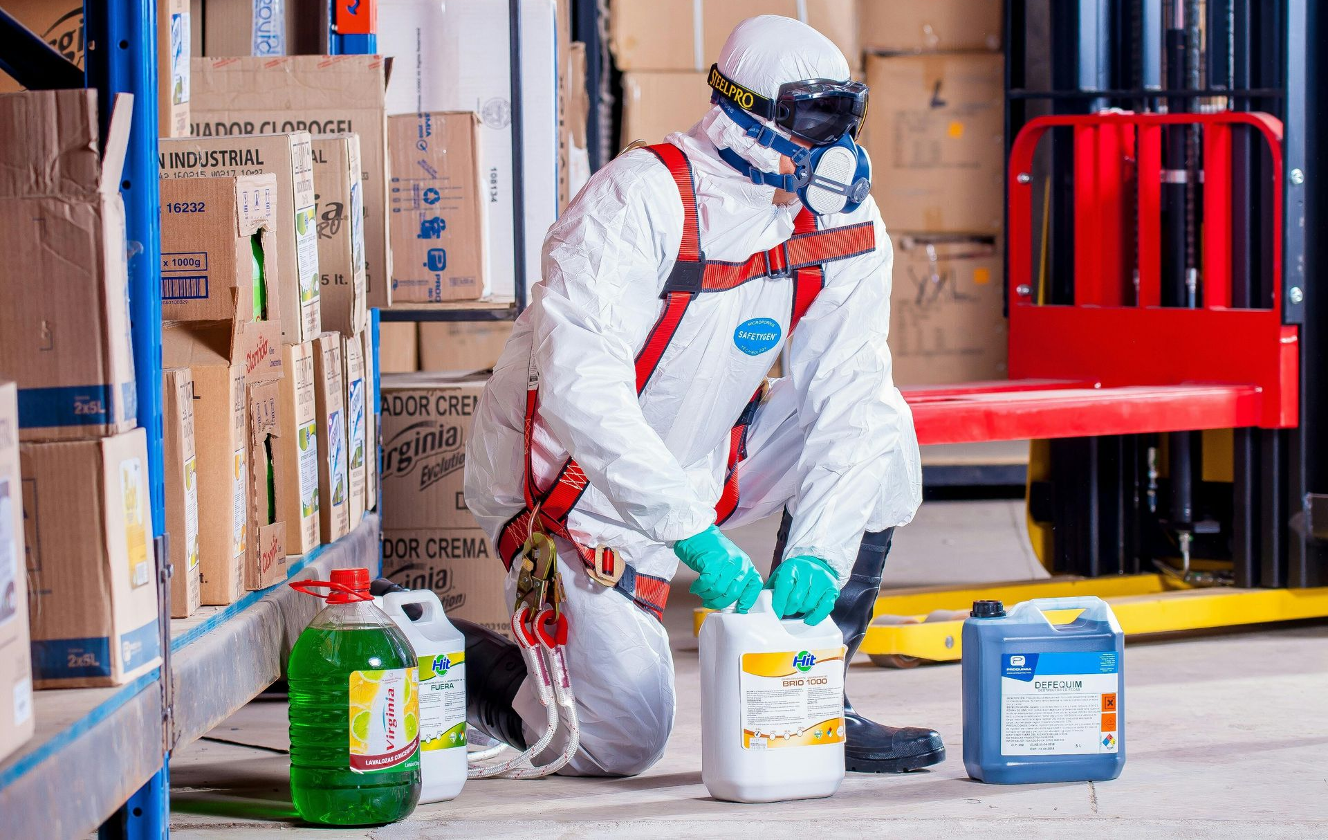 Person in hazmat suit handling chemical container in warehouse.