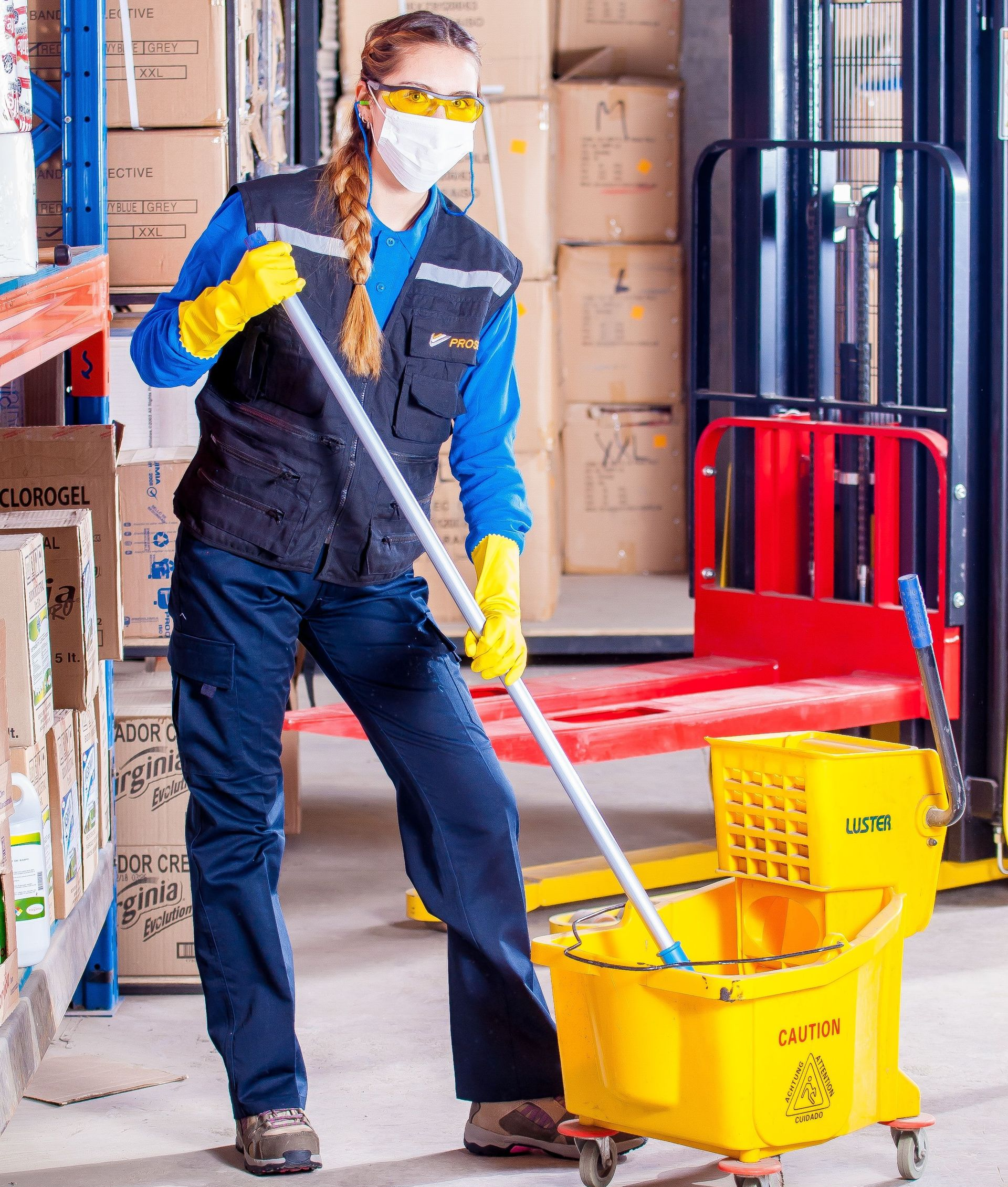 Worker in blue coveralls and yellow gloves mopping a warehouse floor beside a yellow mop bucket