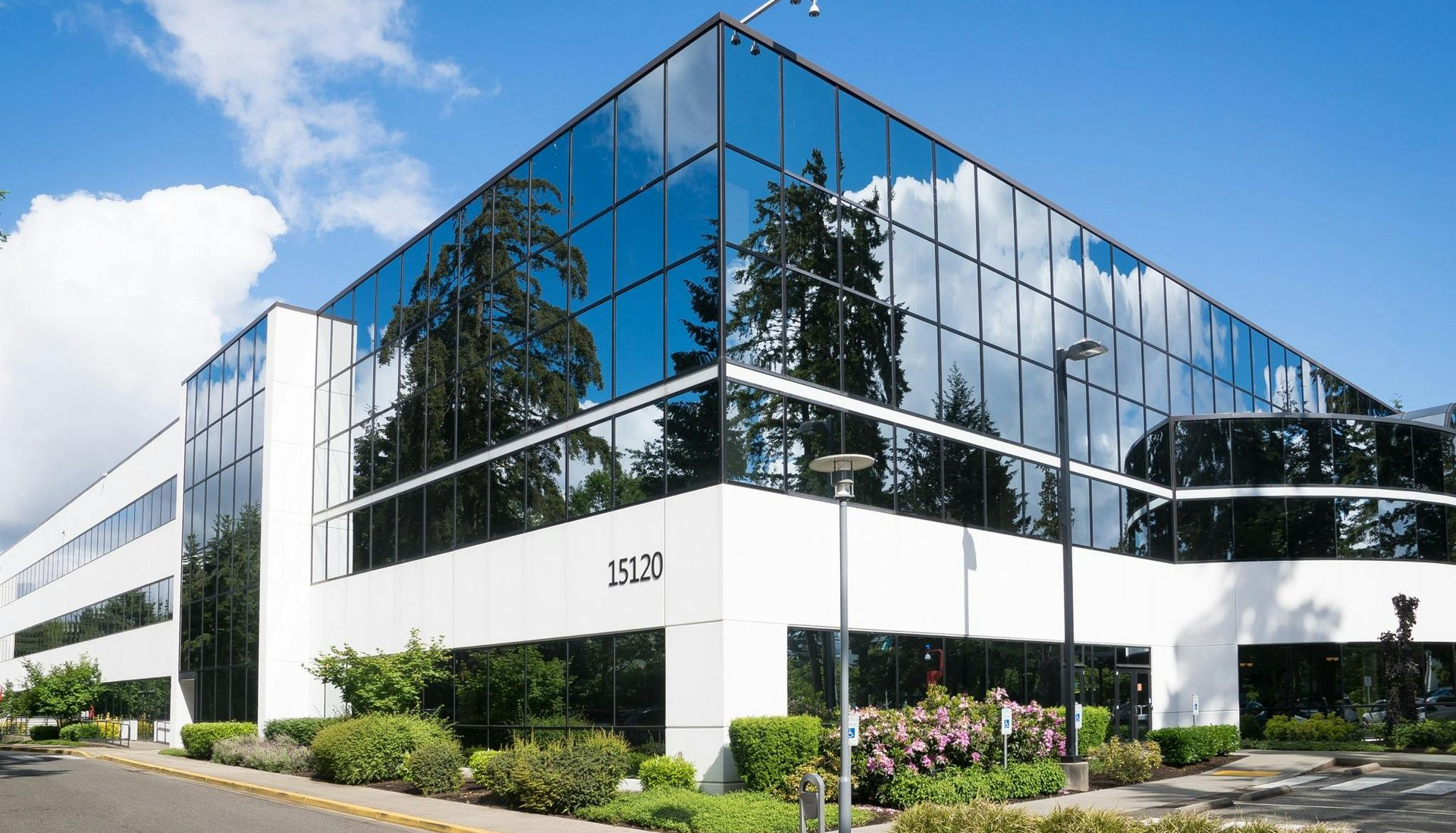 Modern, white office building with large glass windows reflecting trees and blue sky.