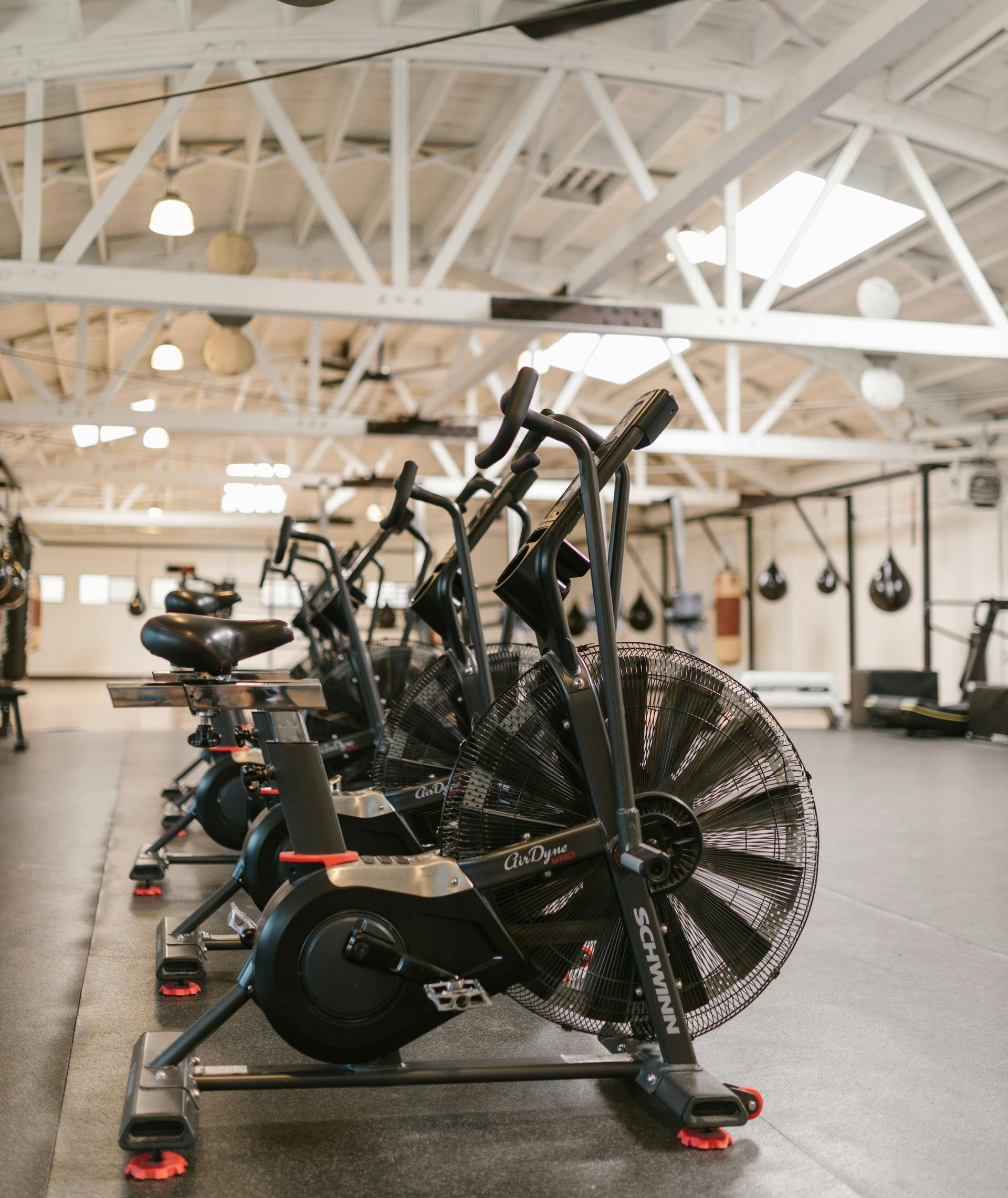 A row of black air bikes with fan-blade wheels inside a gym with high ceilings and white exposed rafters.