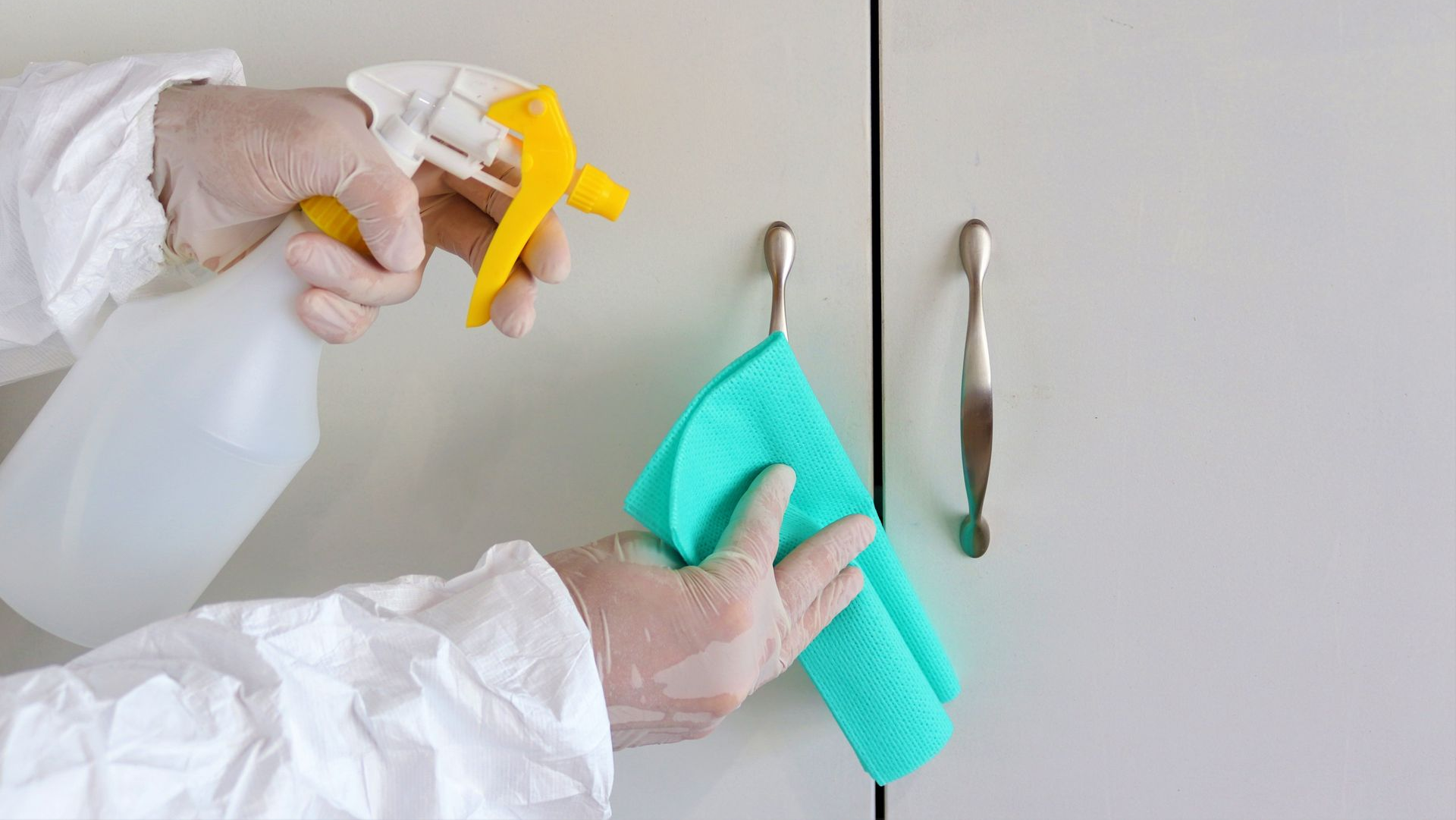 Gloved hands spray and wipe a cabinet with cleaning cloths and spray bottle