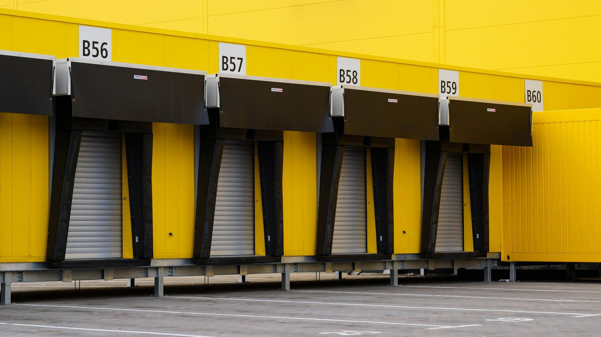 A row of four industrial loading dock bays with metal roller doors against a vibrant yellow wall.