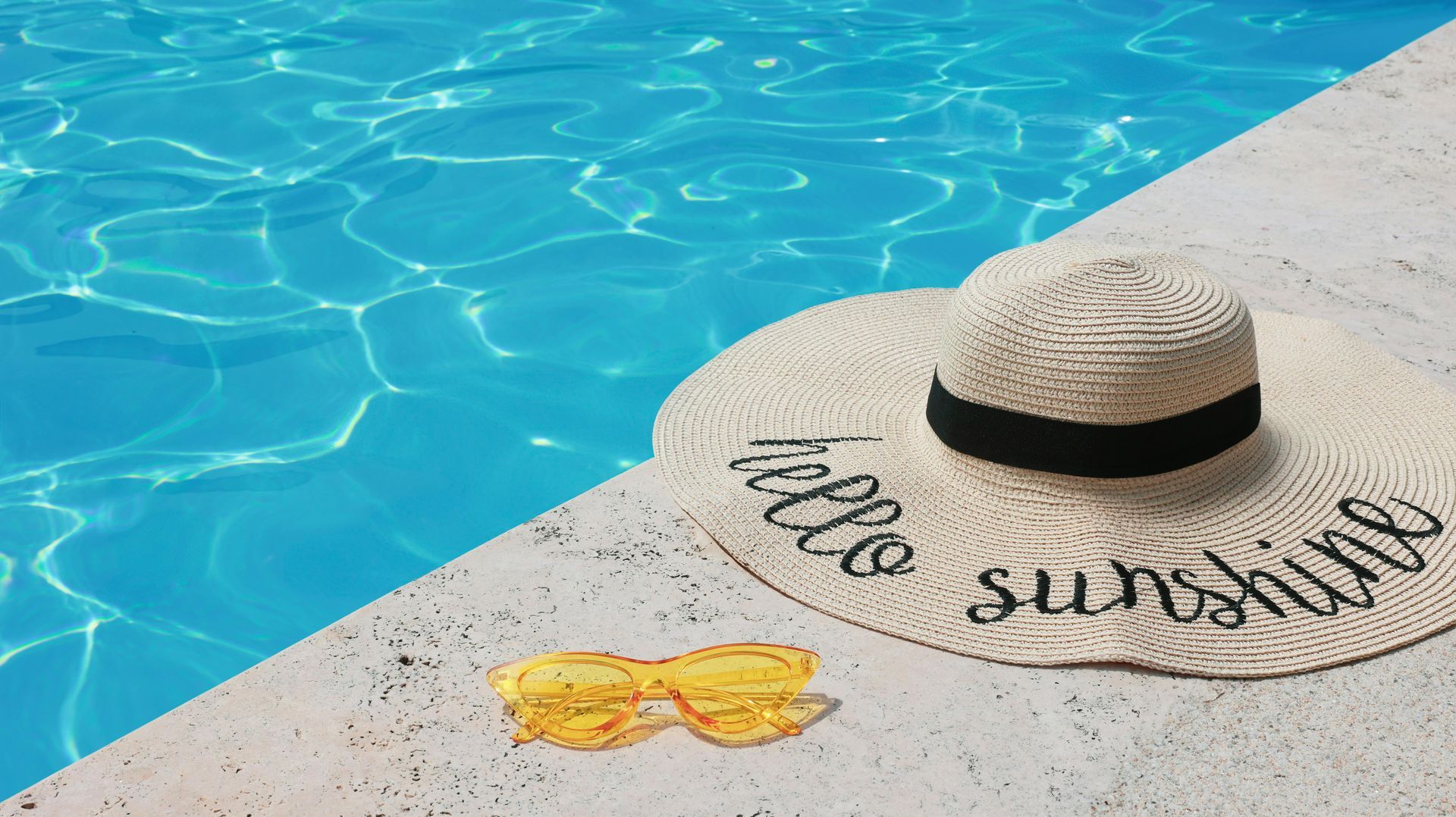 Sun hat and sunglasses on a poolside ledge beside bright blue water