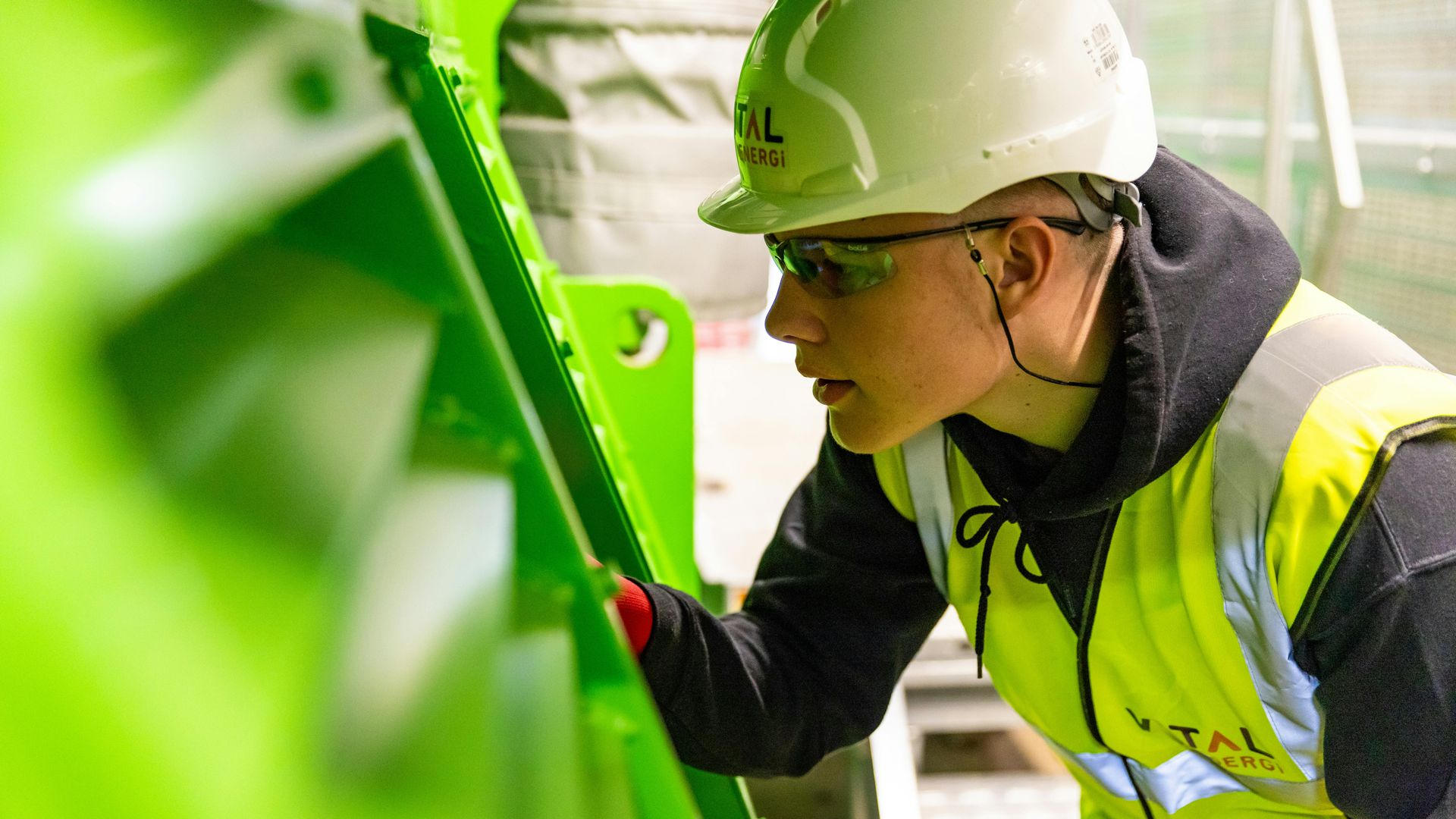 Person in a hard hat and safety vest inspecting machinery, close-up, focusing intently.
