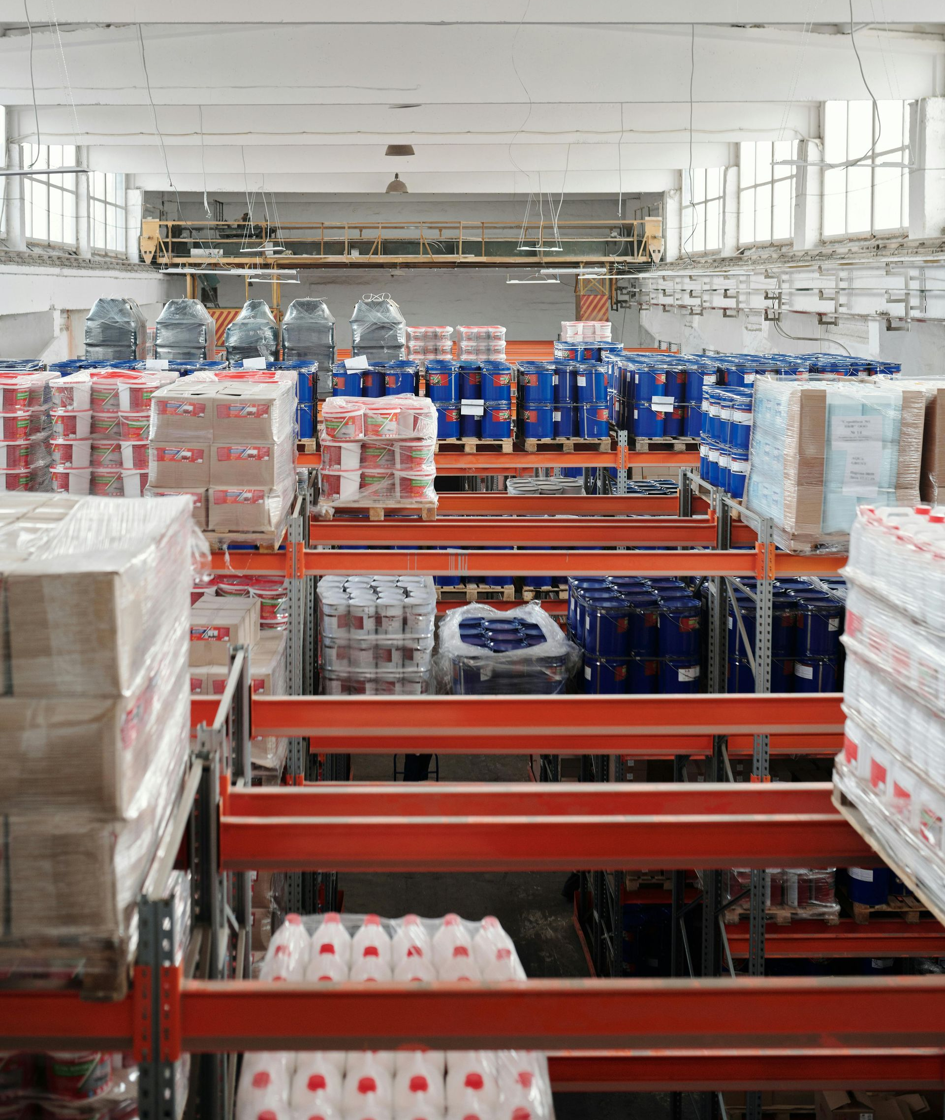 Warehouse interior with rows of tall orange shelving units stocked with stacked, wrapped boxes and blue plastic barrels.