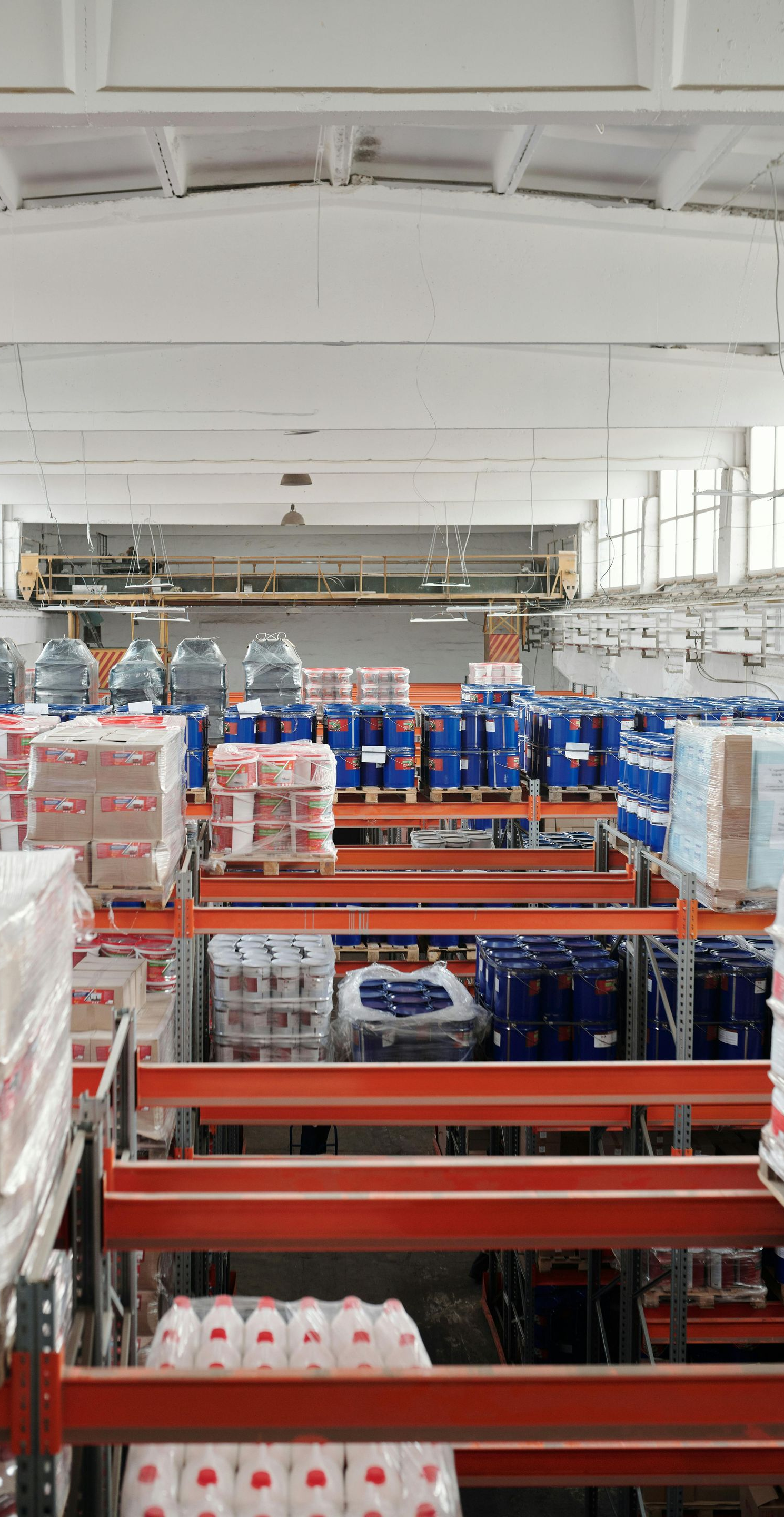 Warehouse interior with stacked goods on orange shelving. Blue barrels, boxes, and white ceiling.