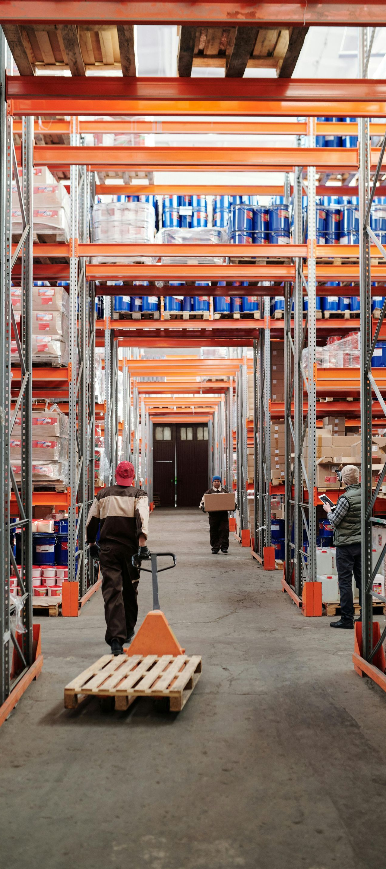 Workers navigate an aisle in a warehouse filled with tall orange metal shelving units containing stacked goods.