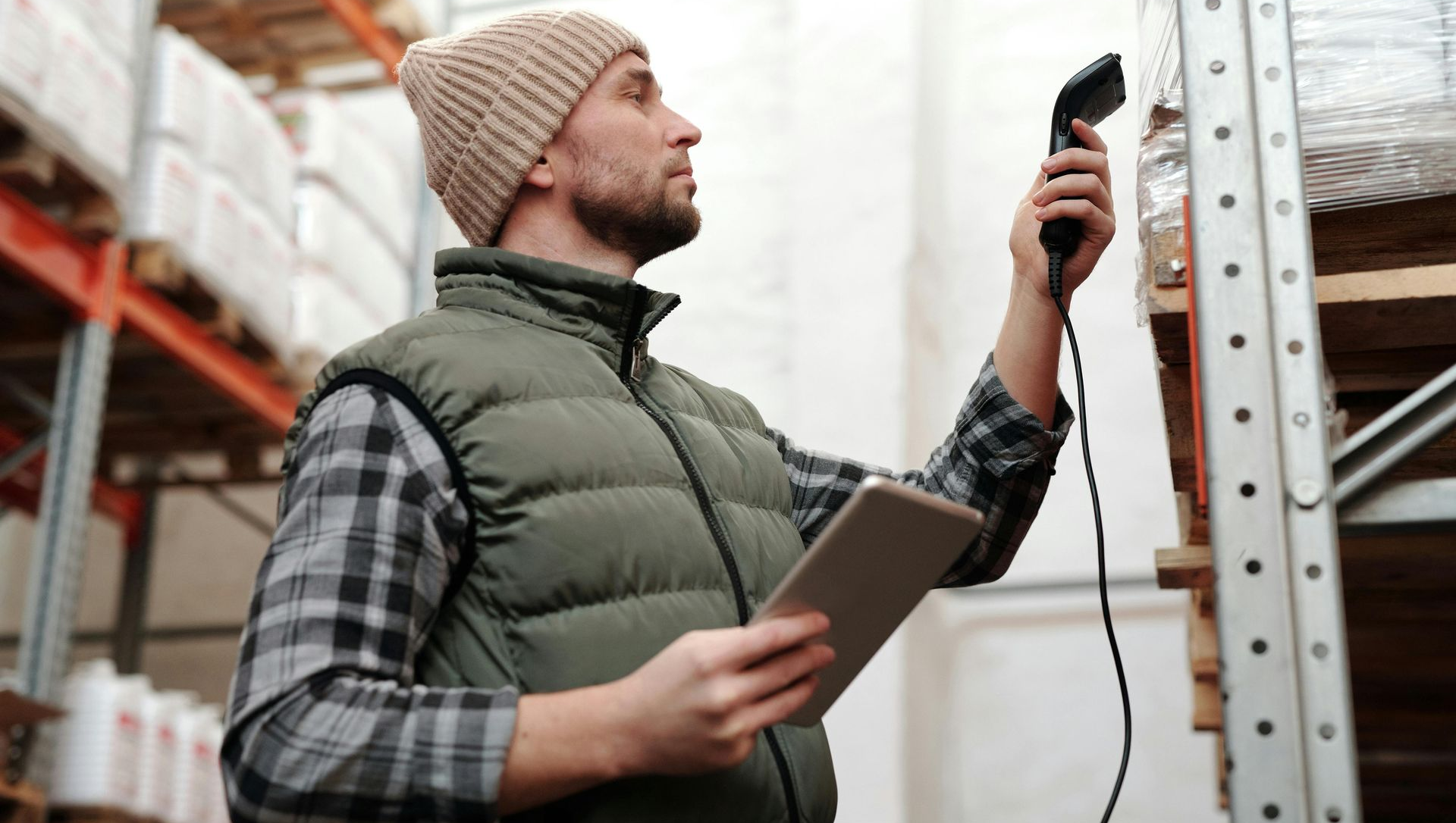 A person in a warehouse wearing a vest and beanie uses a barcode scanner and tablet to track inventory on high shelves.