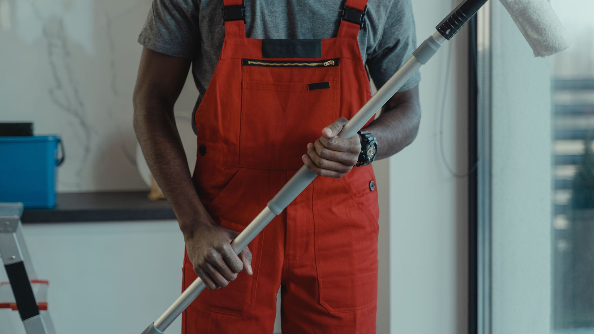 Worker in orange overalls holding a mop in a bright indoor room.