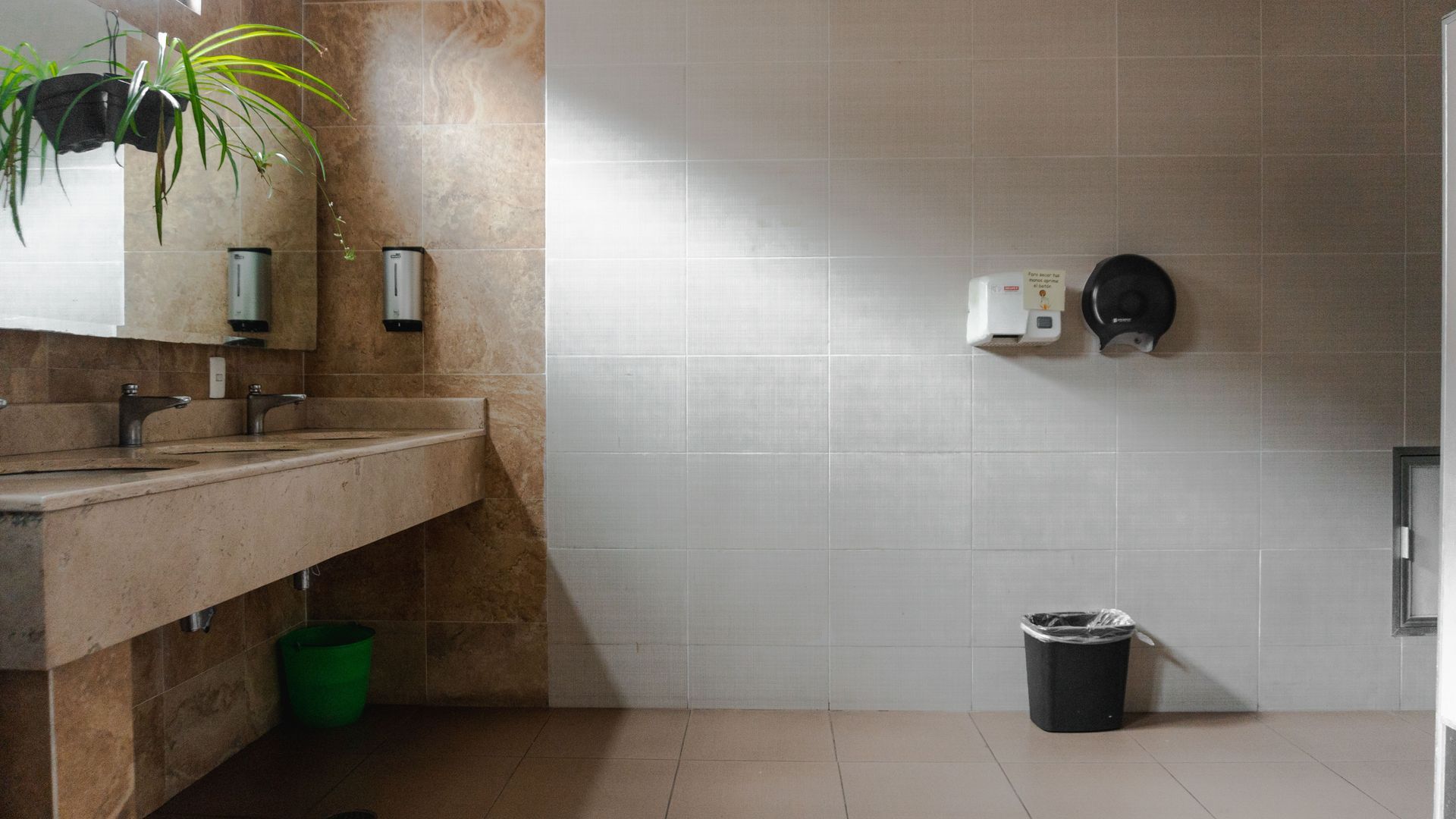 Modern restroom with beige tile walls, concrete sink, wall-mounted soap dispensers, and a trash bin on the floor