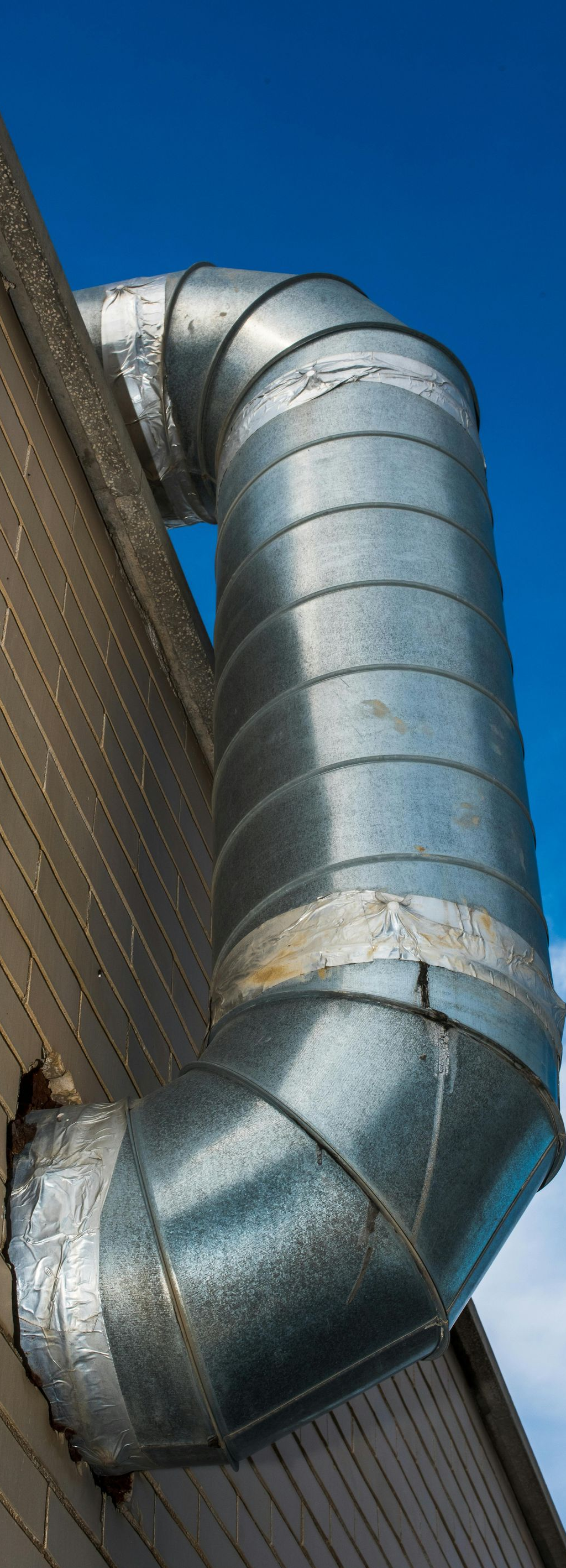 Silver metal ventilation duct on a brick wall against a blue sky.