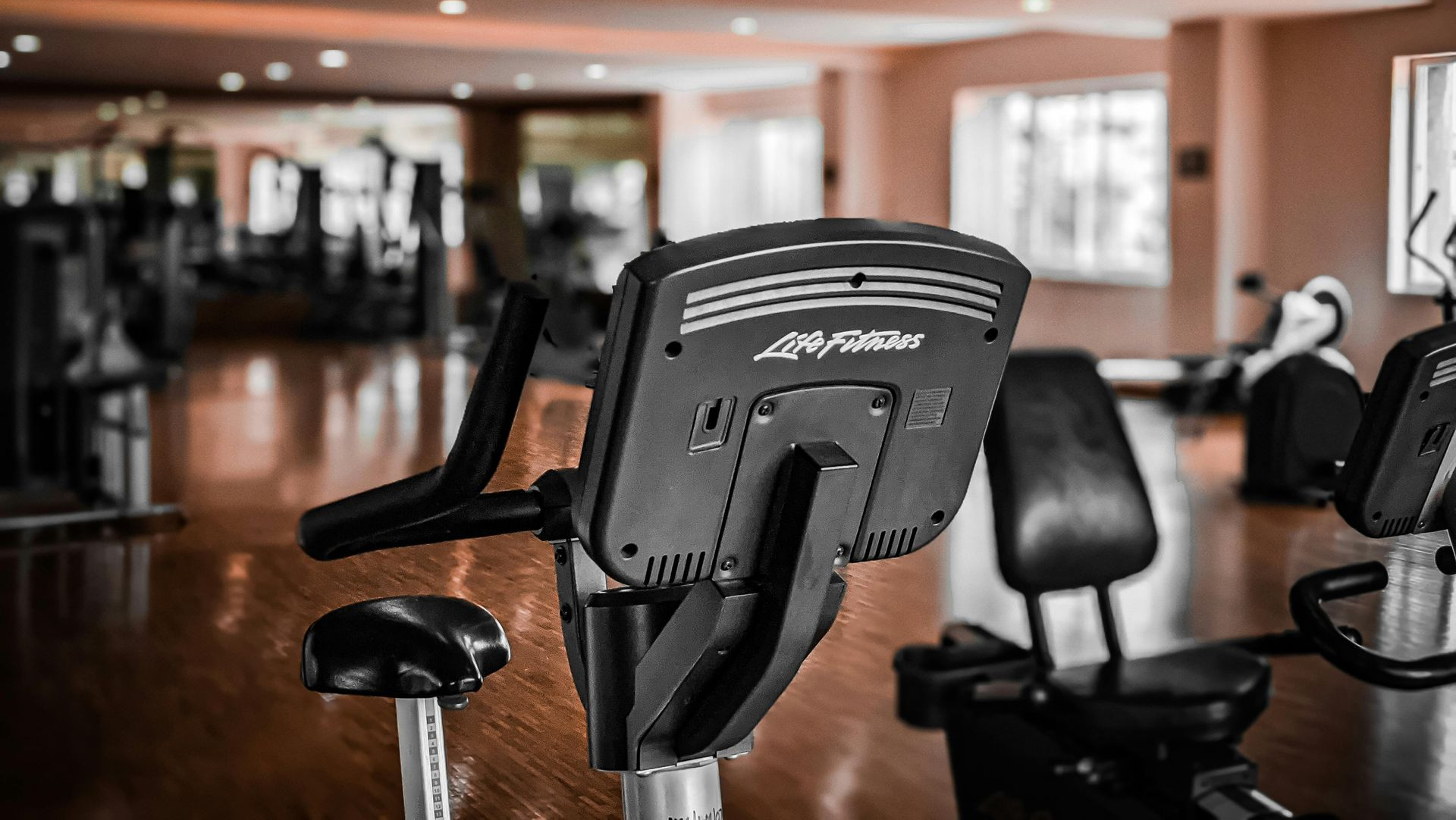 A Life Fitness stationary exercise bike in a well-lit, empty gym with hardwood floors and mirrors in the background.