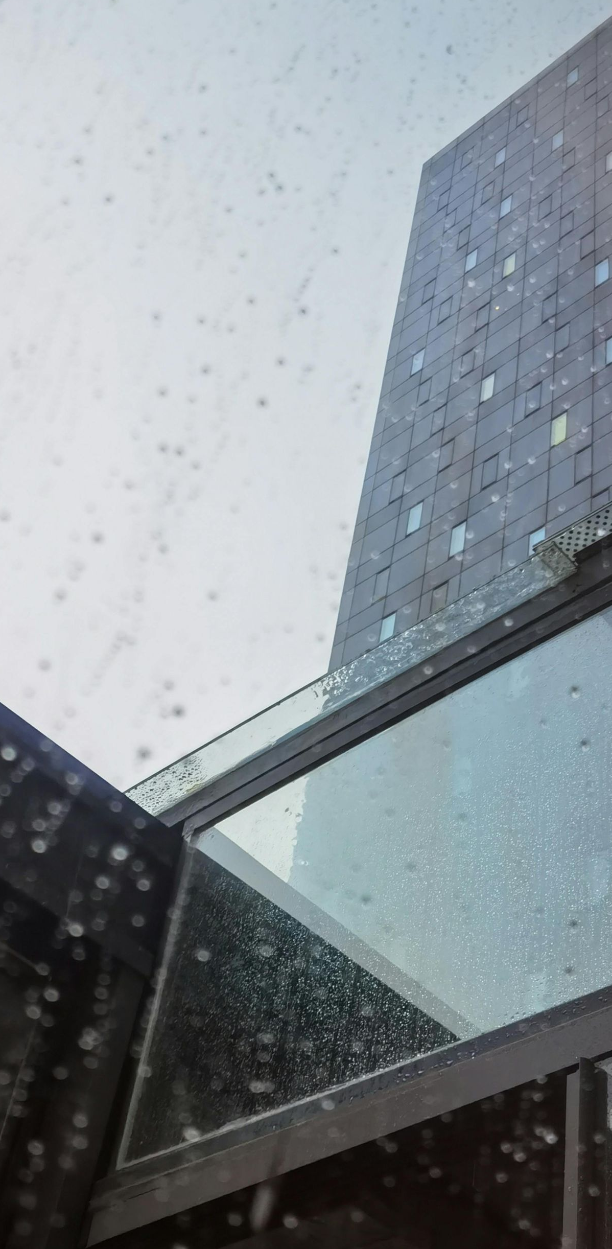 Rain-speckled view of a tall building beside a glass skybridge against a cloudy sky