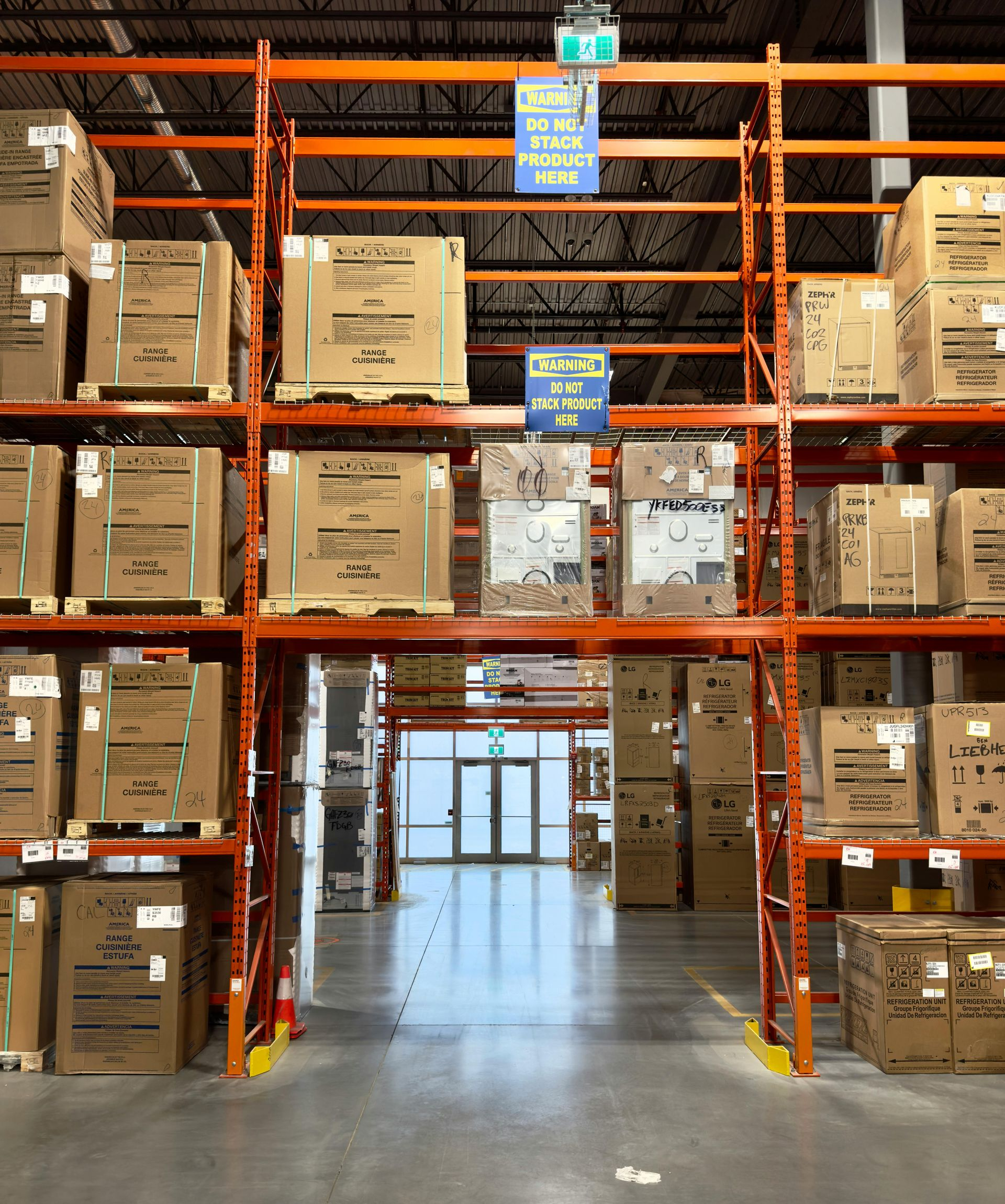 Warehouse aisle with tall orange metal shelves packed with stacked cardboard boxes, leading toward glass exit doors.