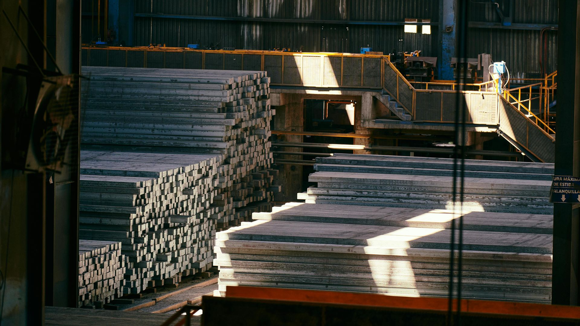 Large stacks of grey metal ingots inside an industrial factory with overhead lighting and a metal walkway.