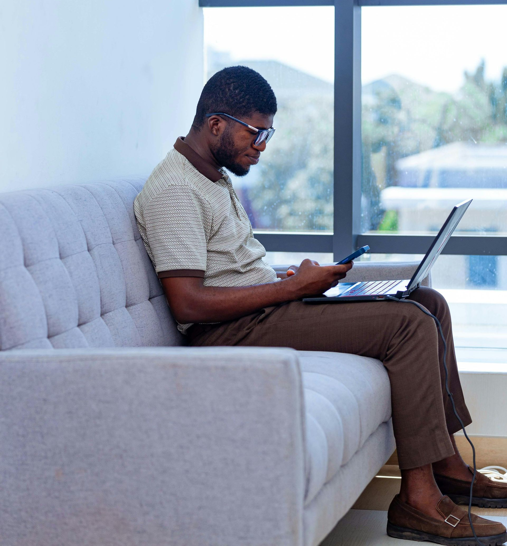 Man using a laptop and phone, seated on a gray couch near a window. He wears glasses, a patterned shirt, and brown pants.
