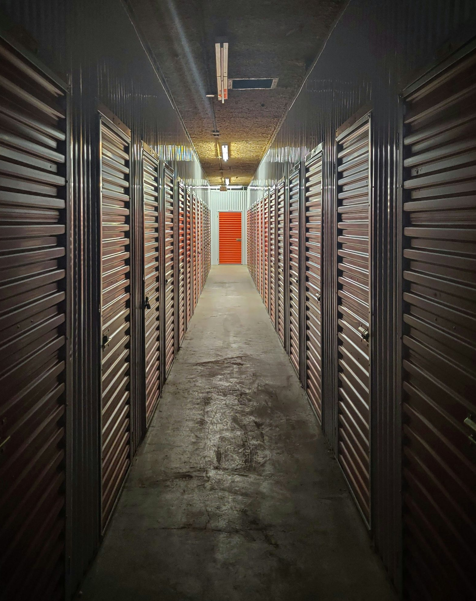 Dim storage unit hallway with brown roll-up doors and a bright orange door at the far end.