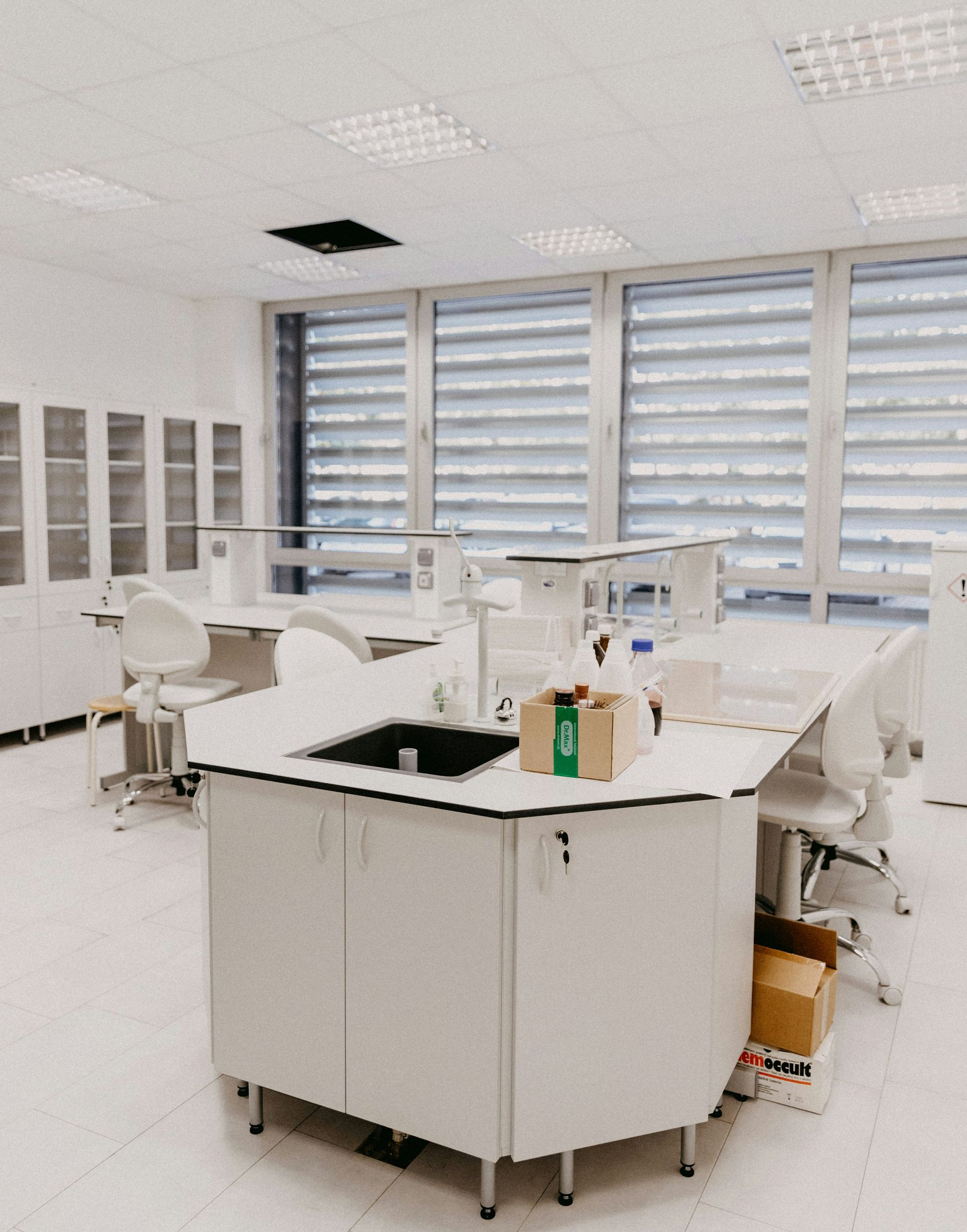 Bright laboratory with white worktables, stools, cabinets, and large windows.