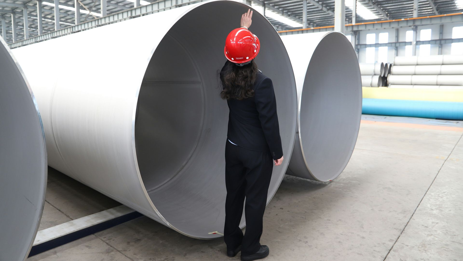 A person in a hard hat inspects the interior of a large, gray industrial pipe inside a factory.