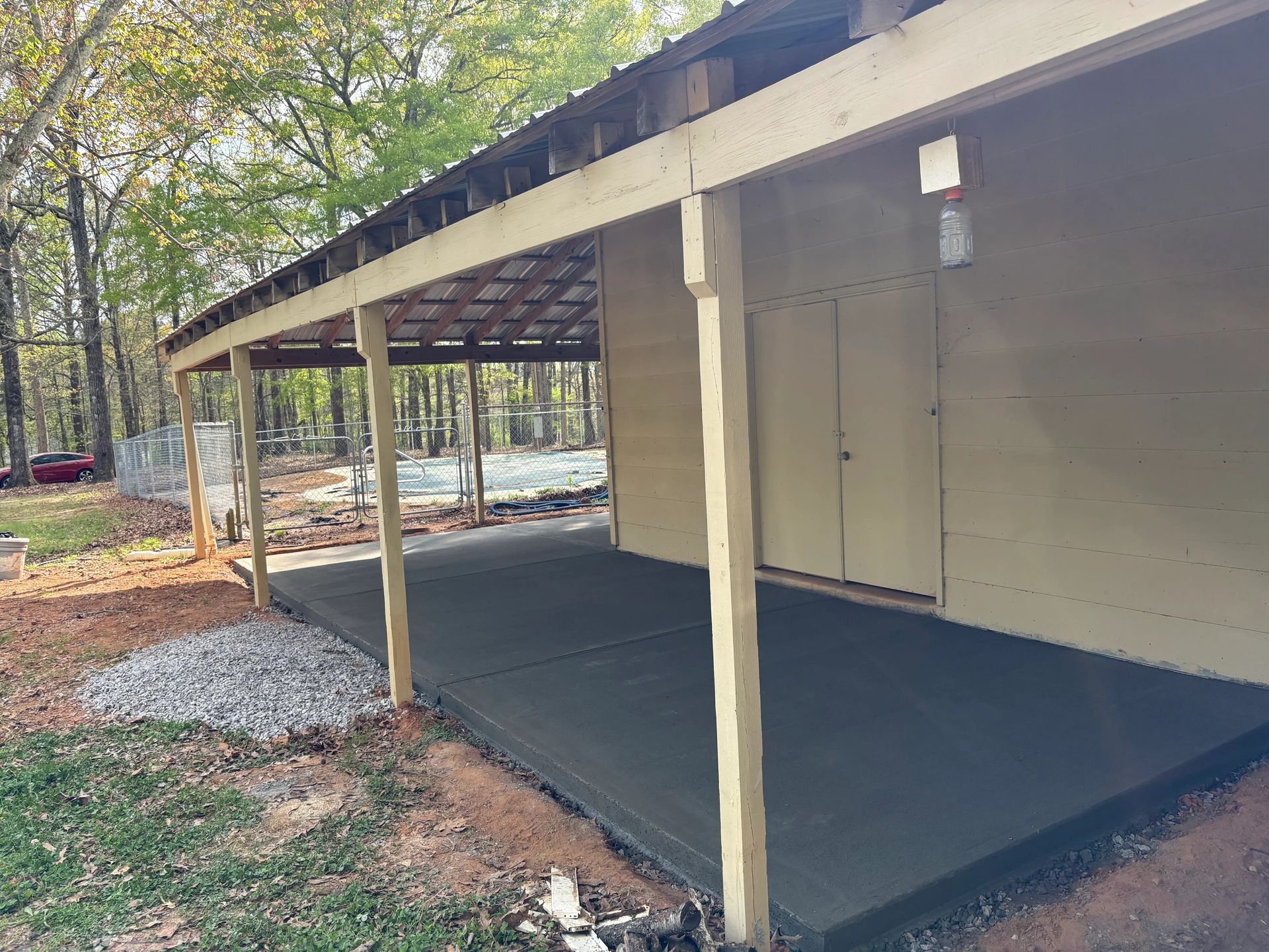 Covered porch with beige posts, dark floor, and a sloped roof beside trees and gravel yard.