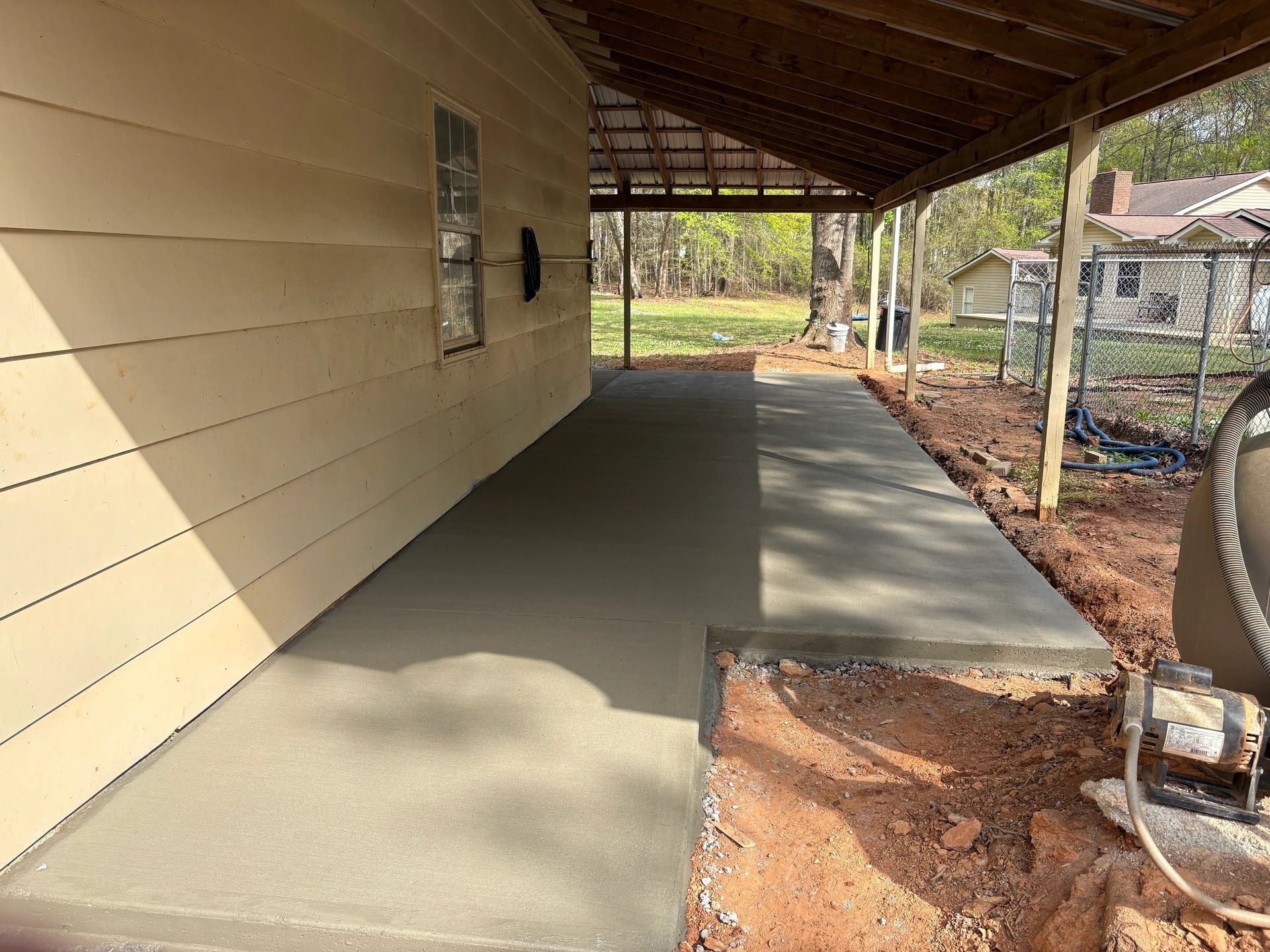 Covered concrete porch beside beige house, with unfinished dirt edge and chain-link fencing nearby