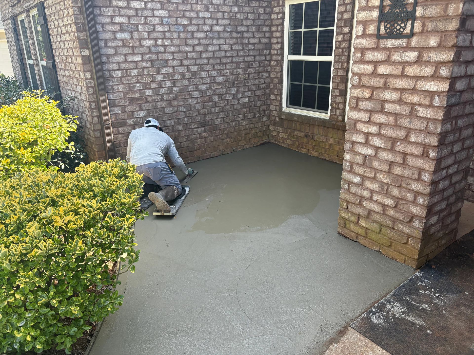Worker smoothing wet concrete beside a brick house and window.