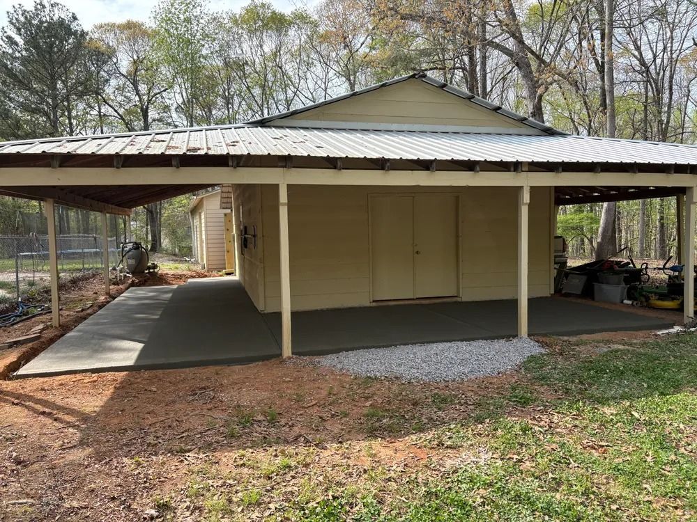 Covered beige carport with a metal roof, concrete pad, and gravel entrance in a wooded yard