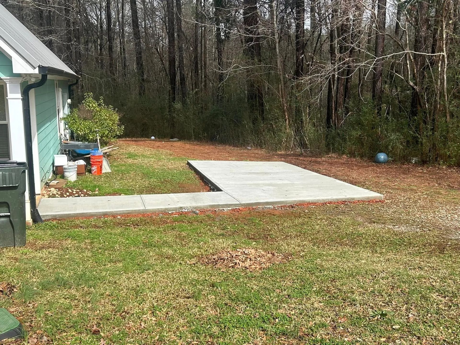 Backyard with a small concrete slab patio beside a house, bordered by grass and woods.