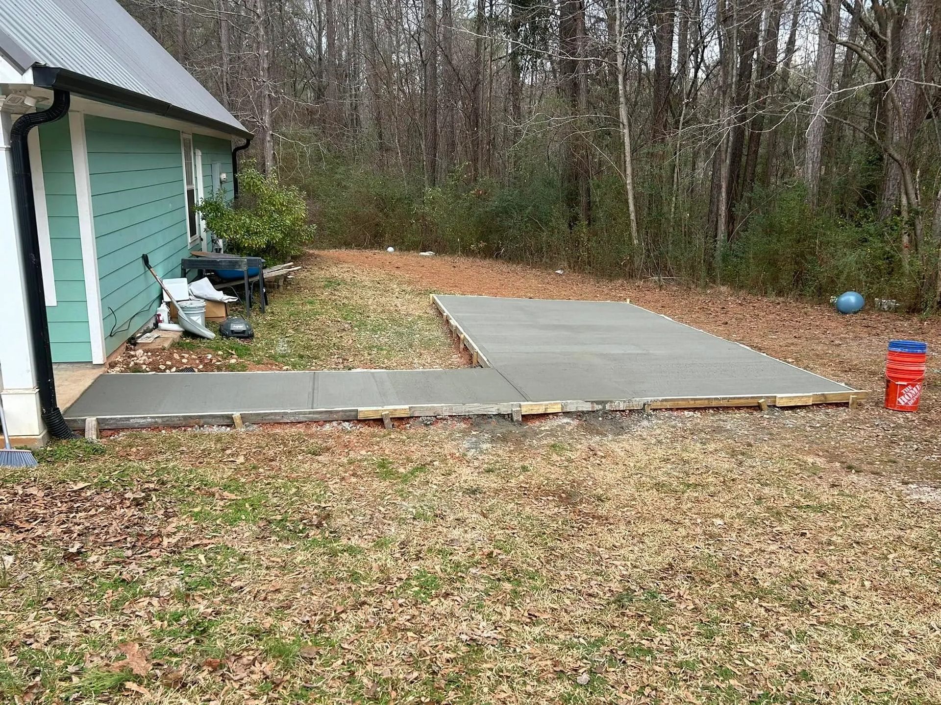 Backyard with a newly poured concrete slab beside a house and wooded trees in the background