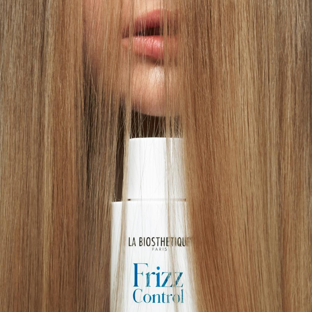 Le visage d'une femme se cache derrière une bouteille de produit anti-frisottis.