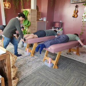 Therapist stretching three clients on massage tables in a cozy studio with plants and violin decor