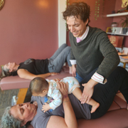 Two adults and a baby in a therapy room, with one adult supporting the baby on another adult’s legs.