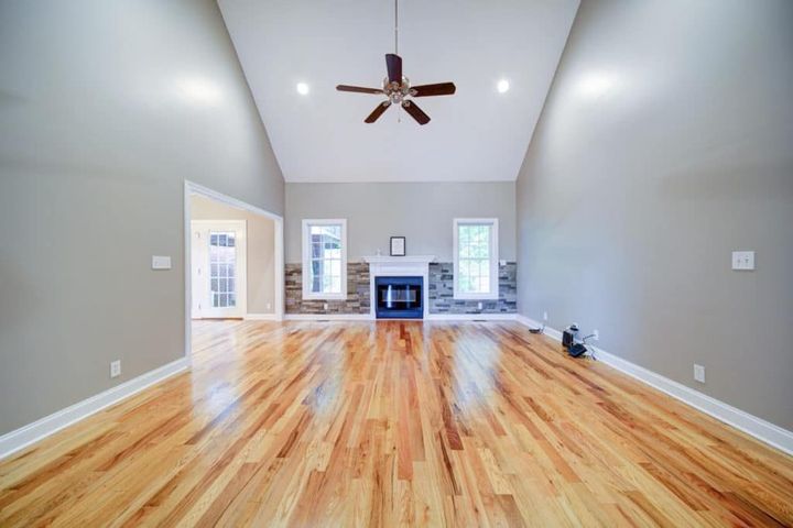 Empty living room with wood floor, high ceiling, fireplace, and two windows.