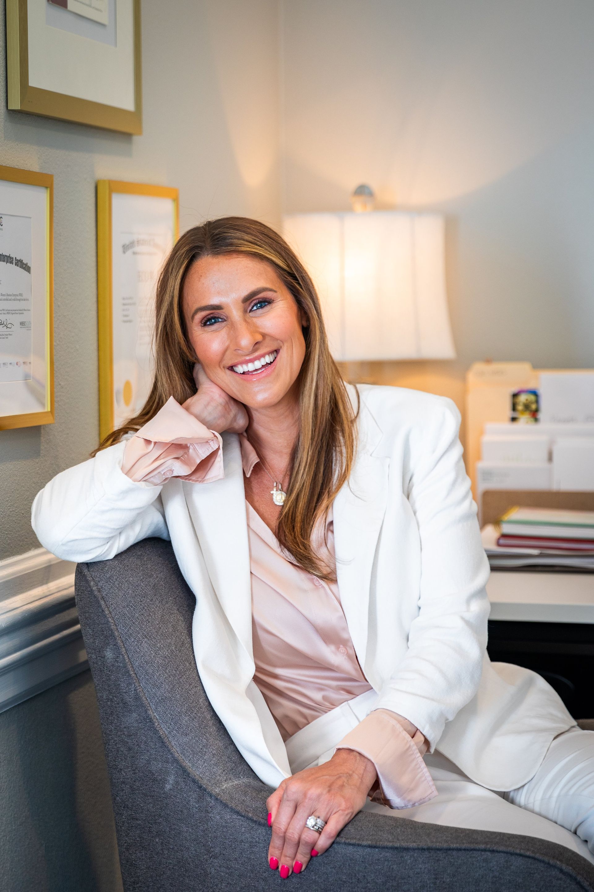 A woman in a white suit is sitting in a chair and smiling.