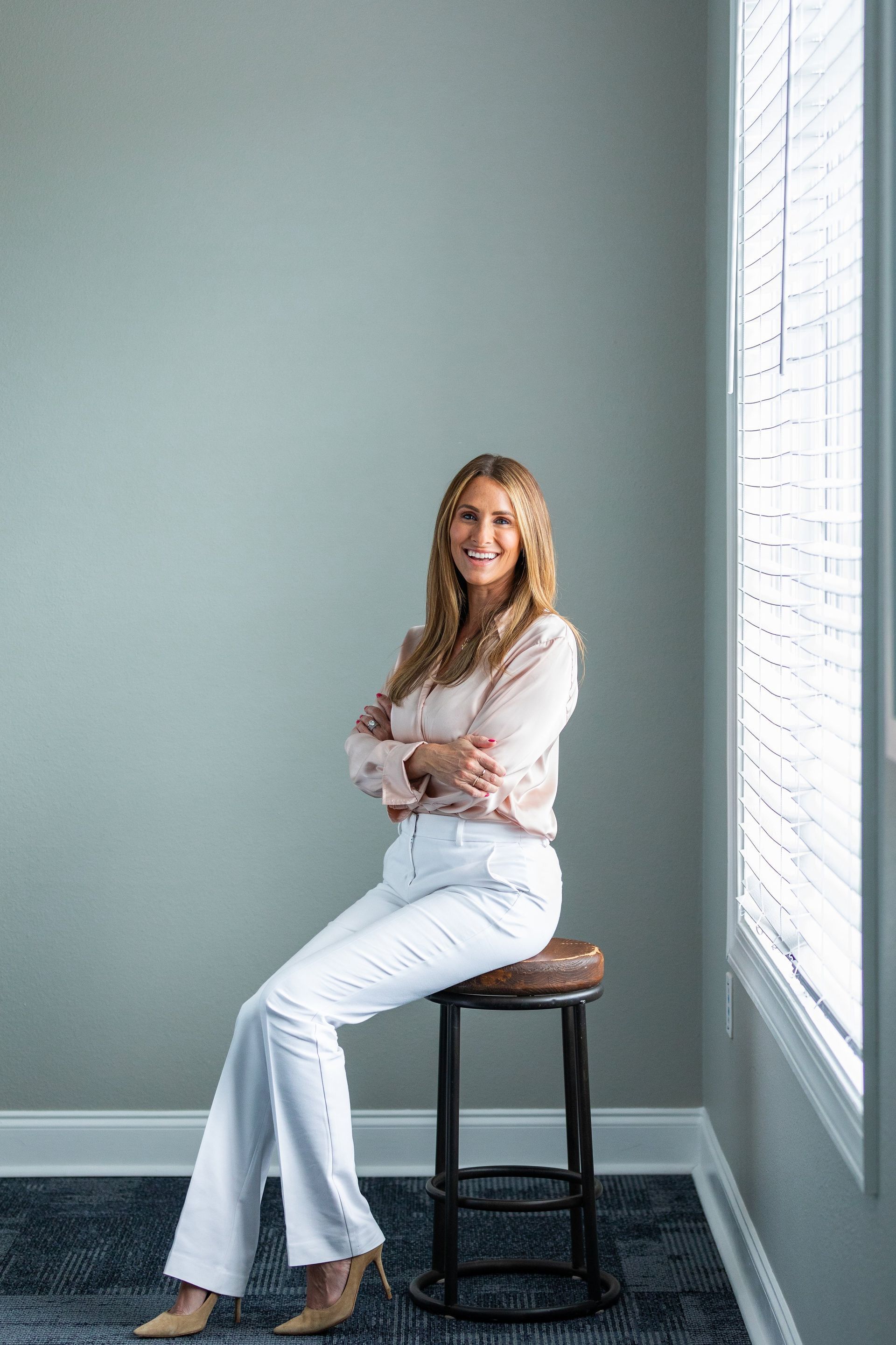 A woman is sitting on a stool in front of a window.