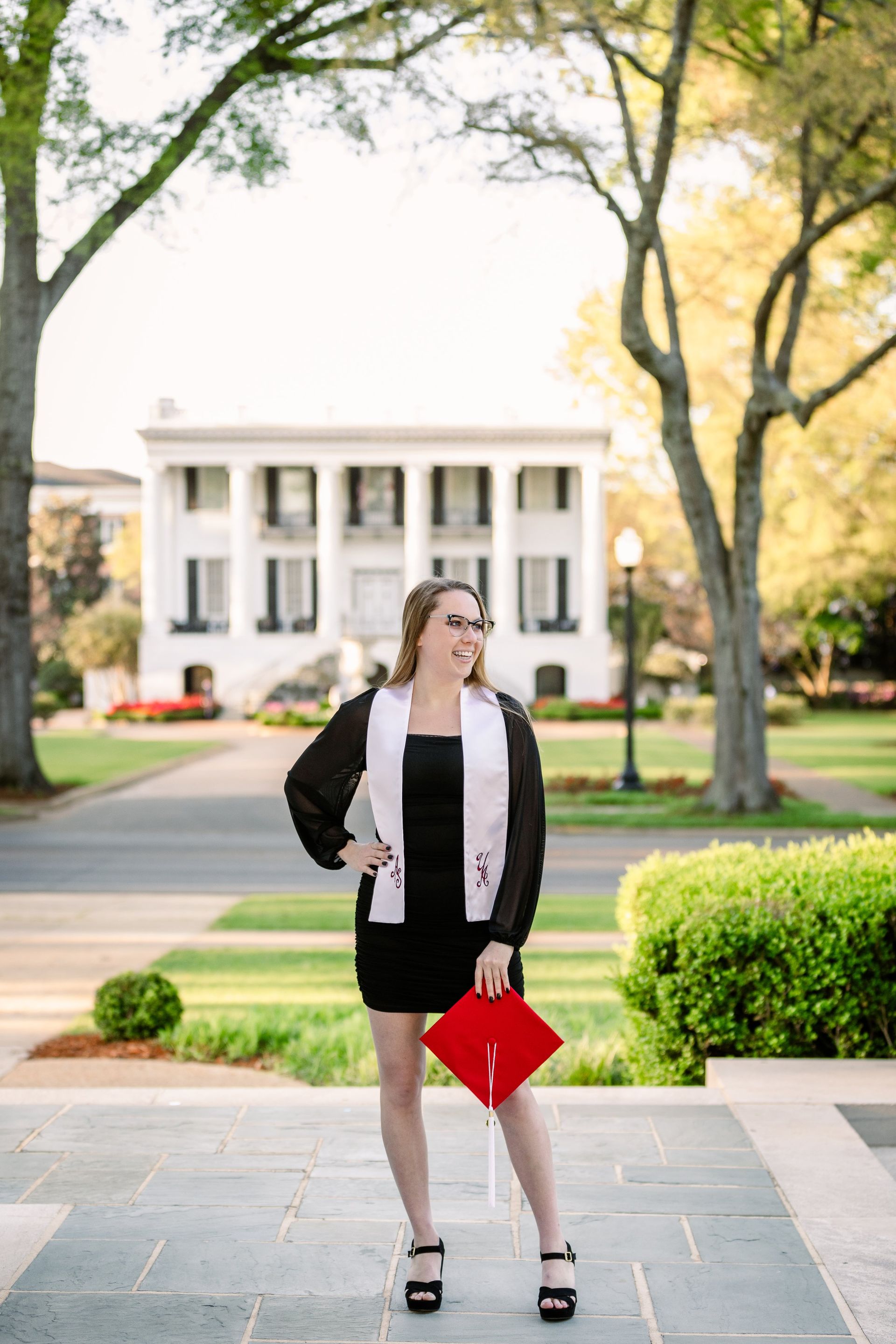A woman in a black dress and red cap and gown is standing in front of a white building.