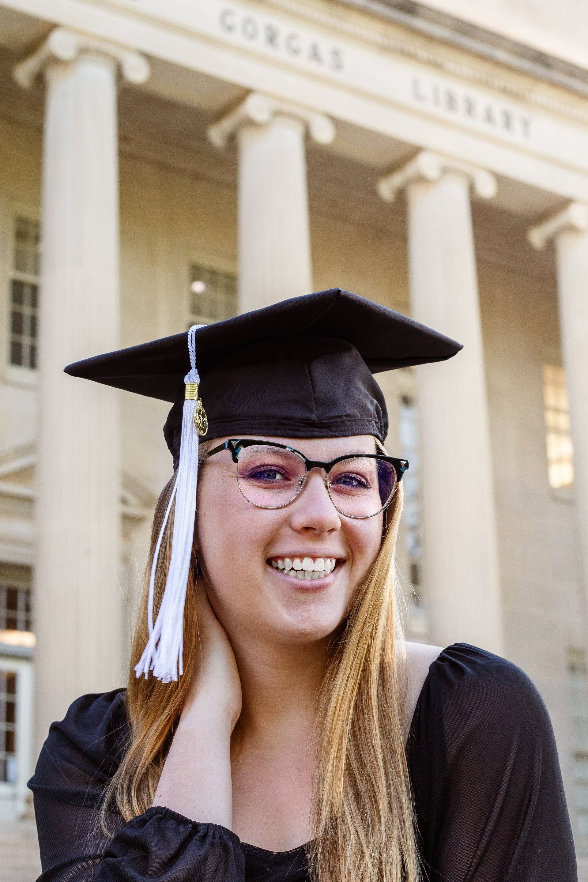 A woman wearing a graduation cap and gown is smiling in front of a library.