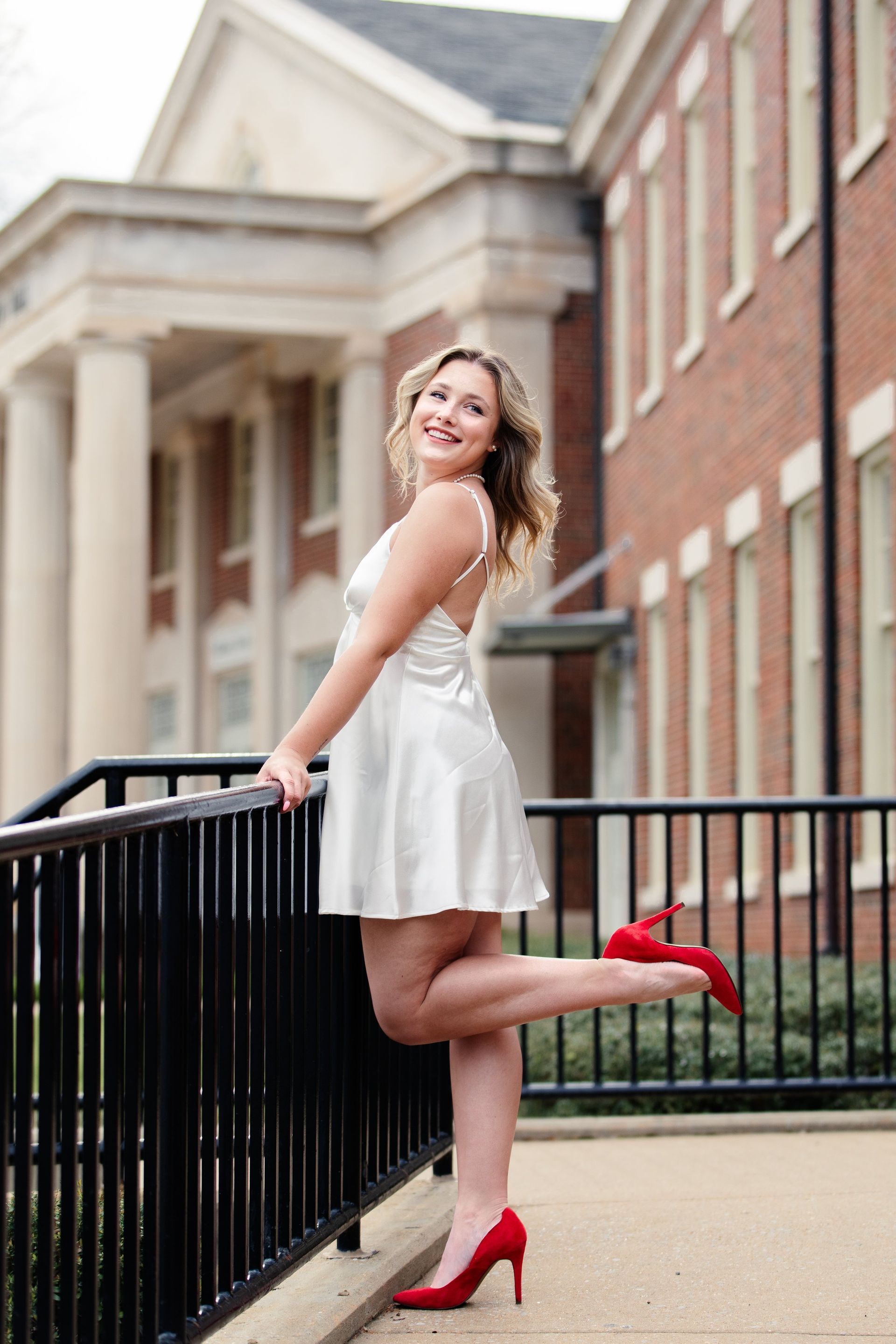 A woman in a white dress and red heels is leaning on a railing.