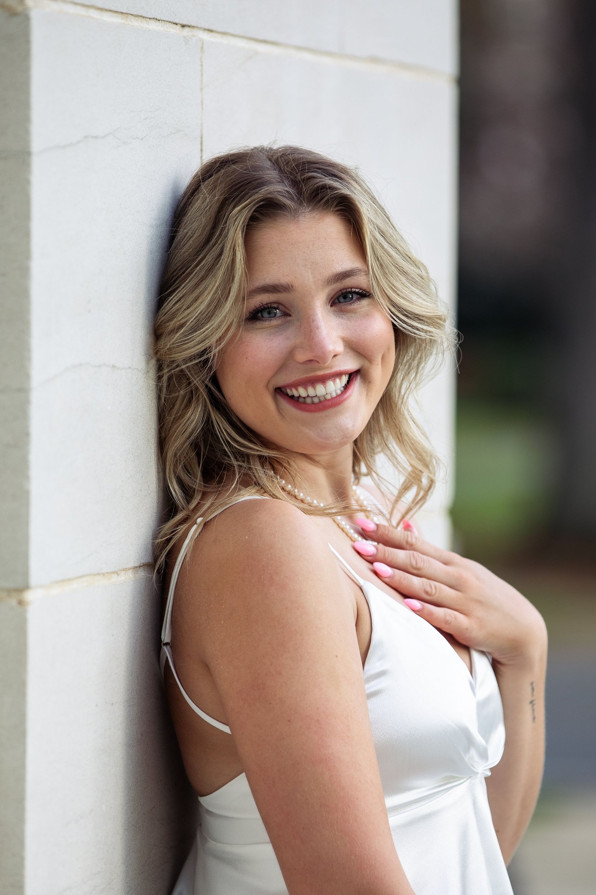 A woman in a white dress is leaning against a wall and smiling.