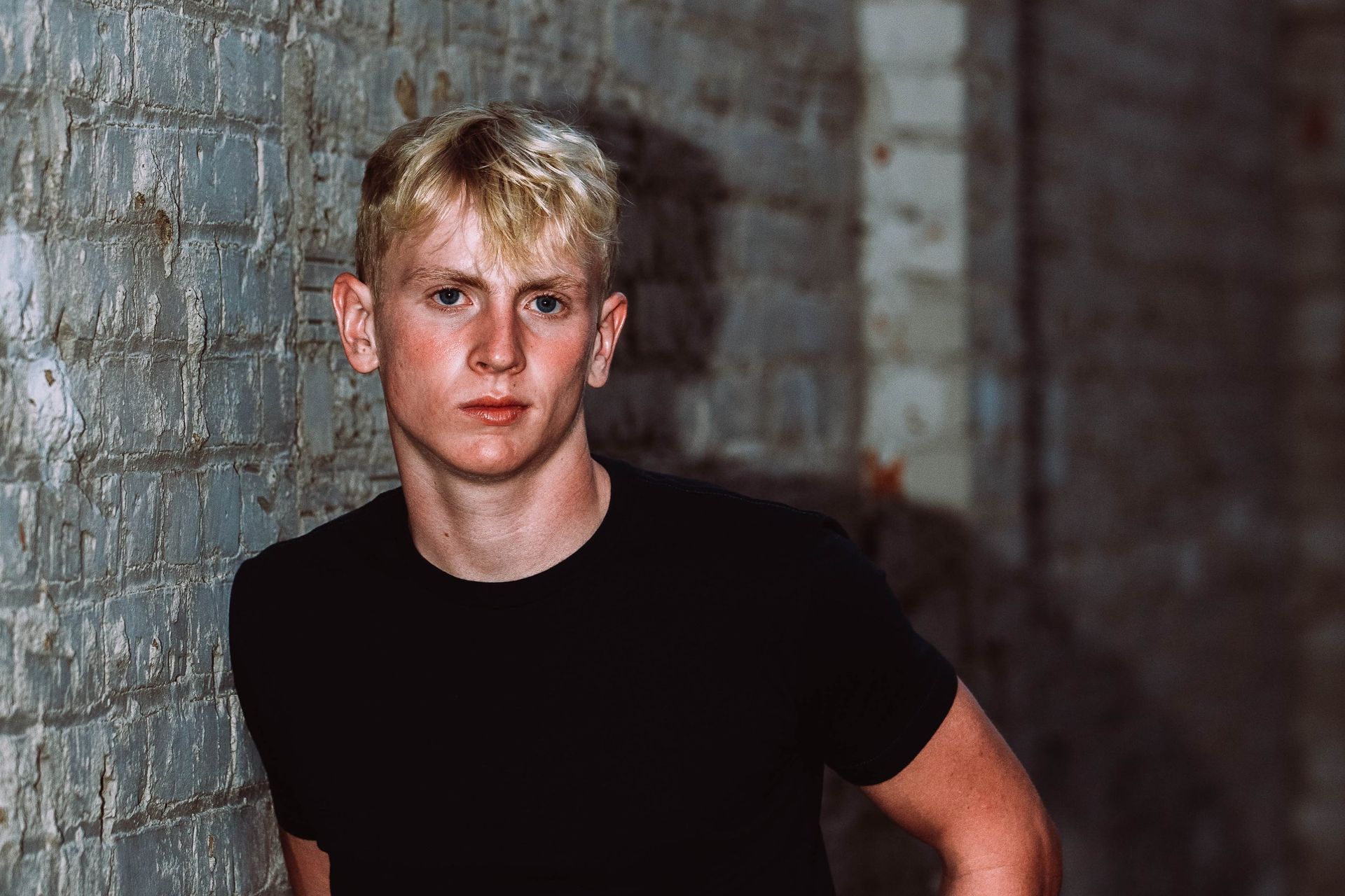 A young man is leaning against a brick wall wearing a black shirt.