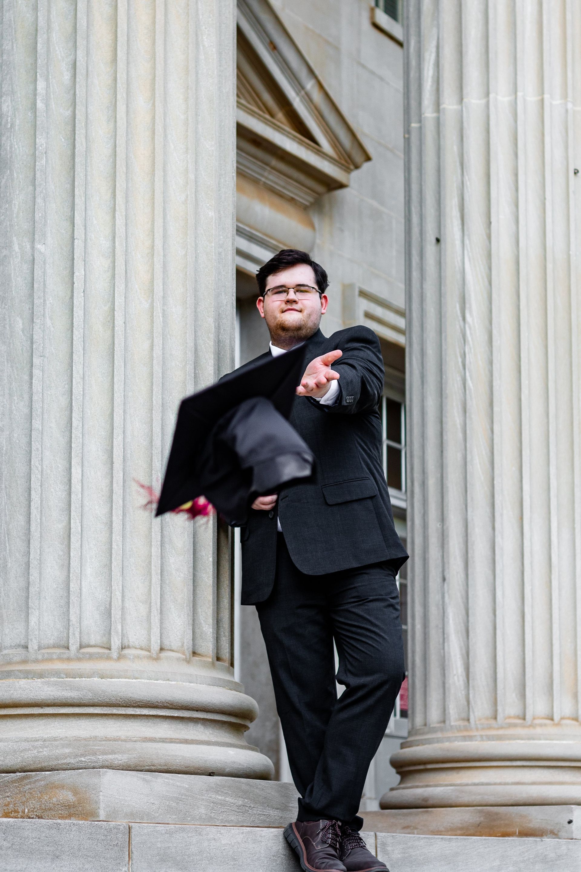 A man in a suit is standing in front of a building holding a graduation cap.