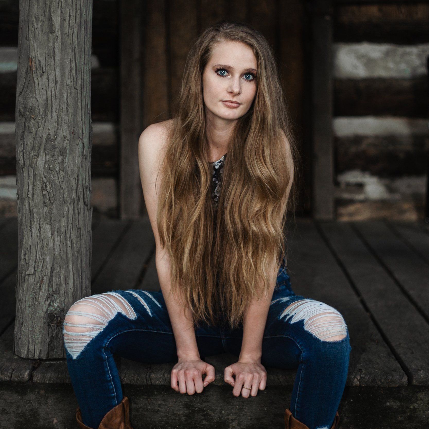 A woman with long hair is sitting on a wooden deck next to a tree.