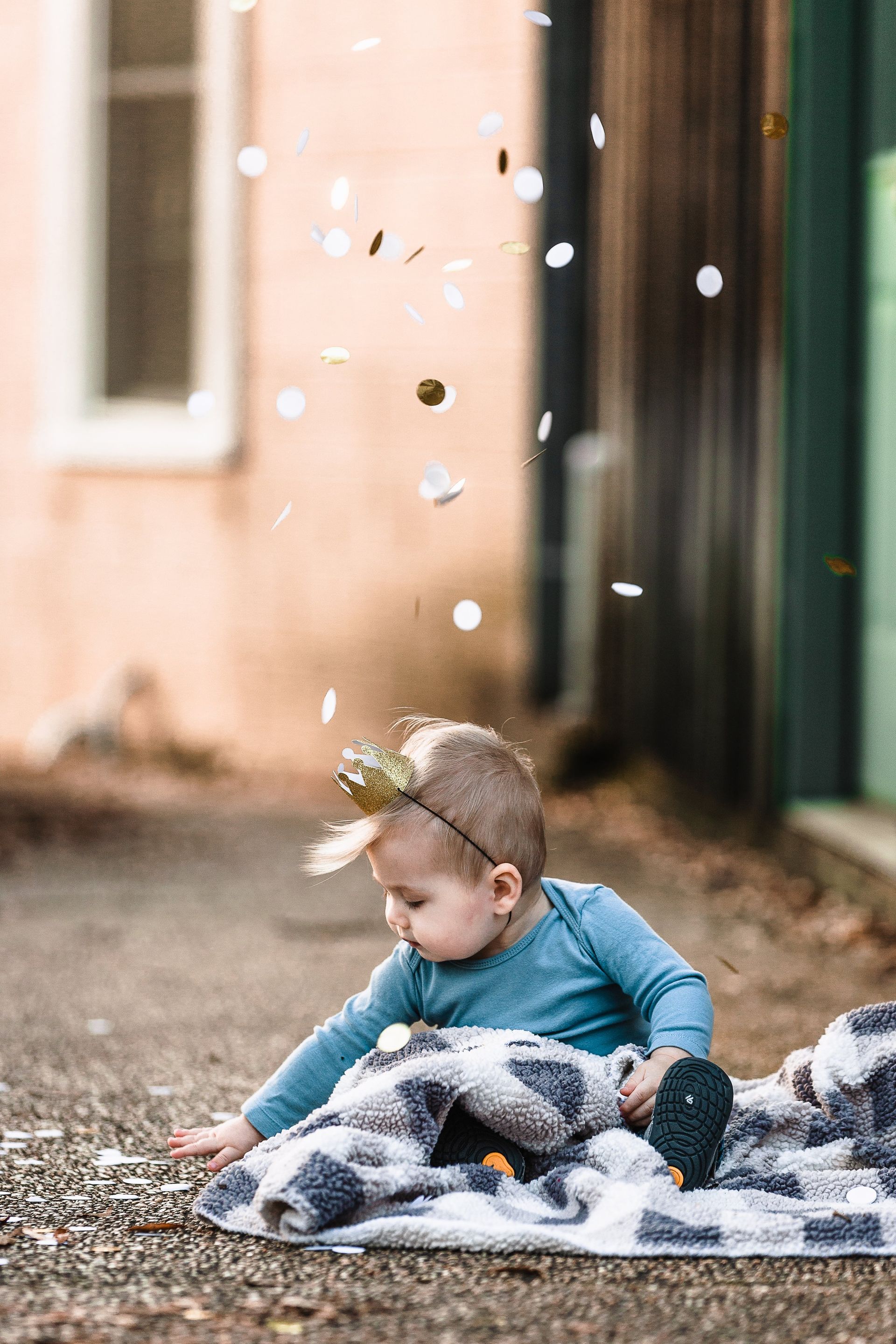 A baby is sitting on a blanket on the ground with confetti falling around her.