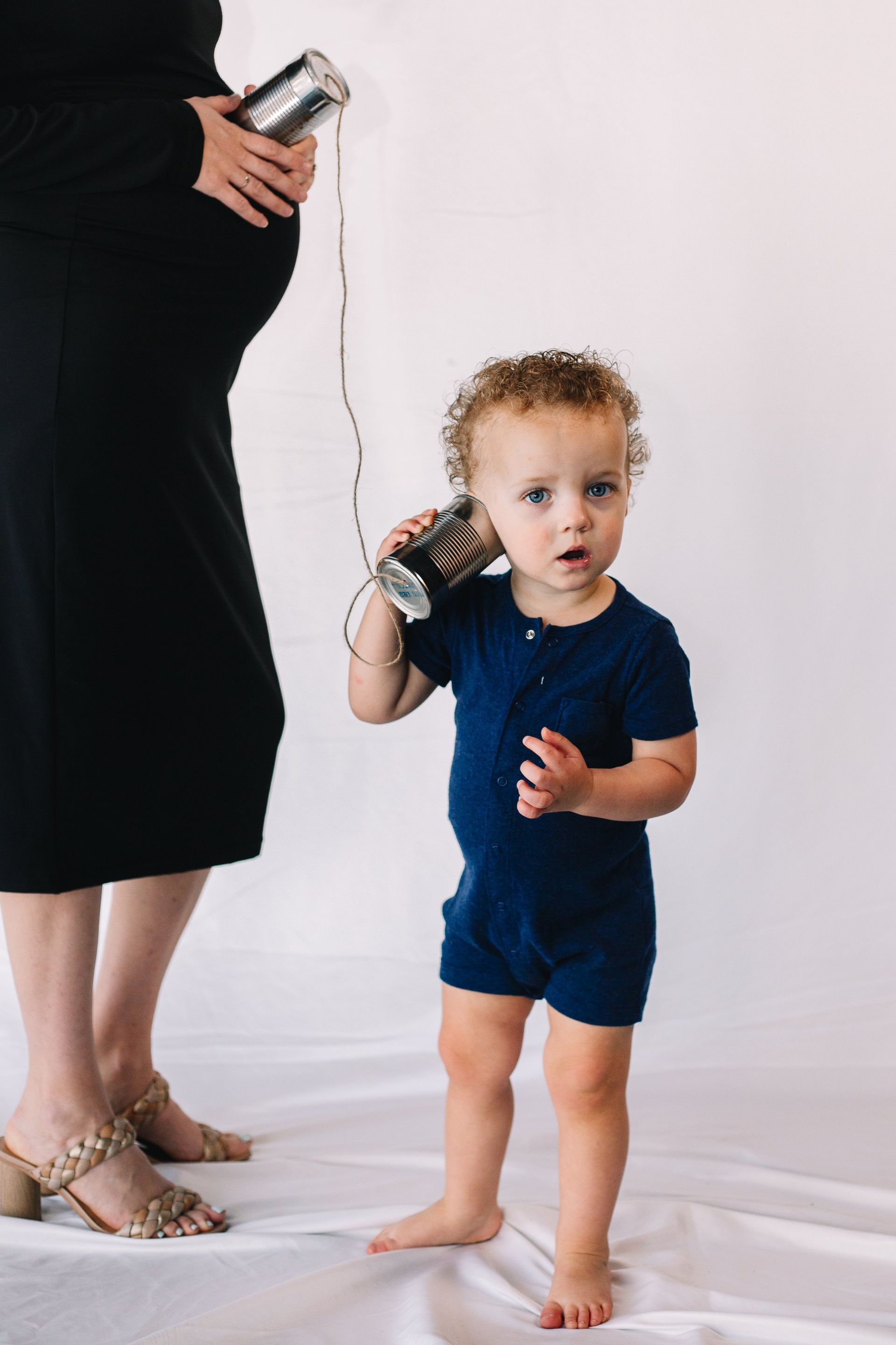 A baby is talking on a tin can phone next to a pregnant woman.