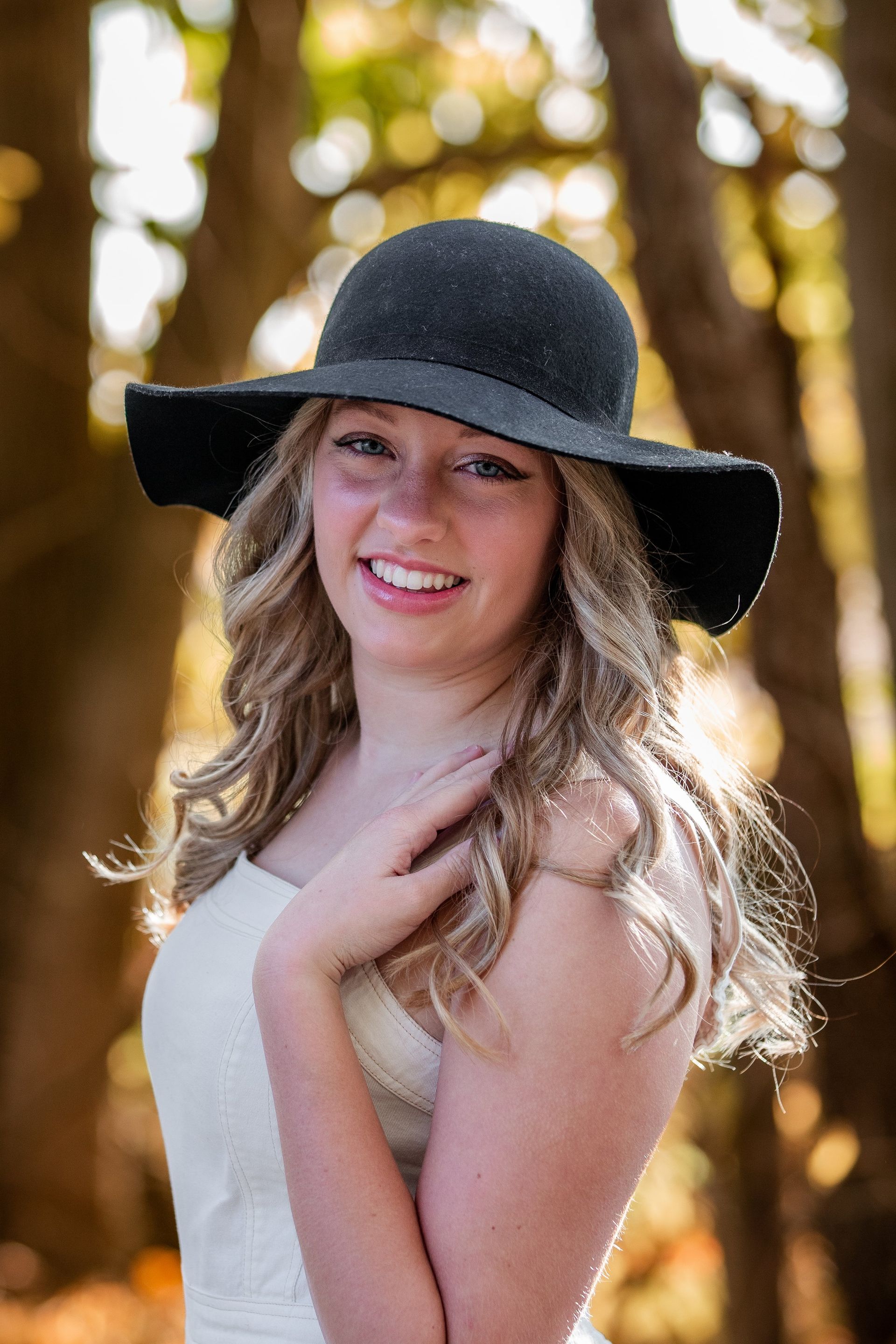 A young woman wearing a black hat and a white dress is standing in the woods.