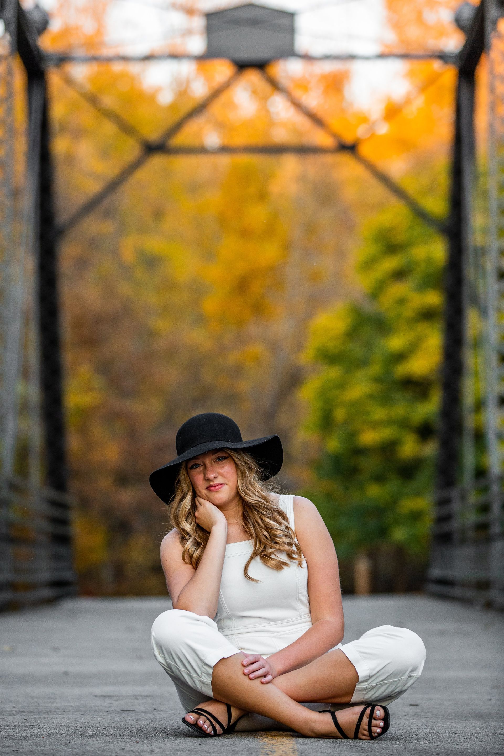 A woman in a hat is sitting on the ground in front of a bridge.
