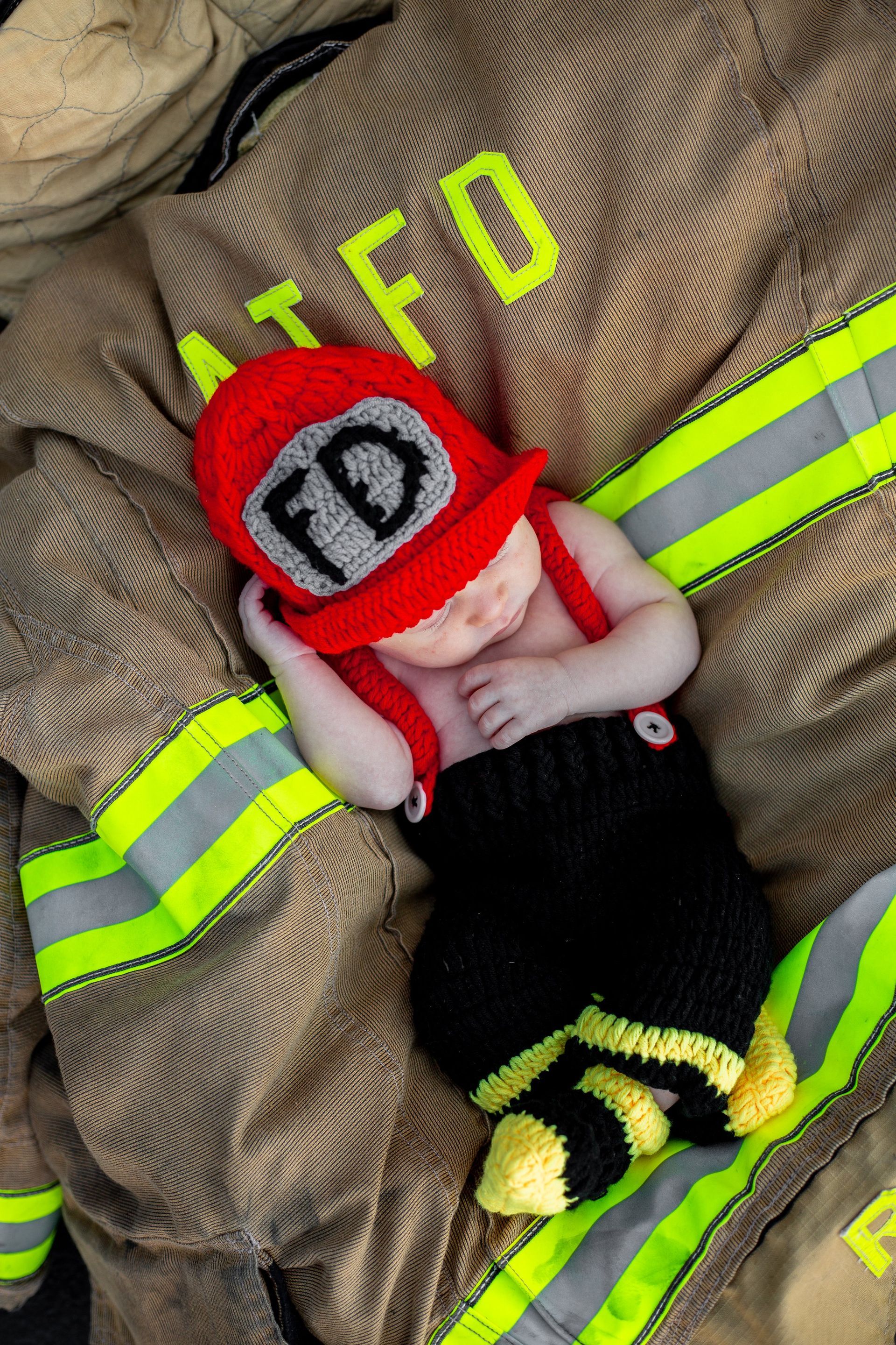 A baby in a fireman costume is sleeping on a fireman 's jacket.