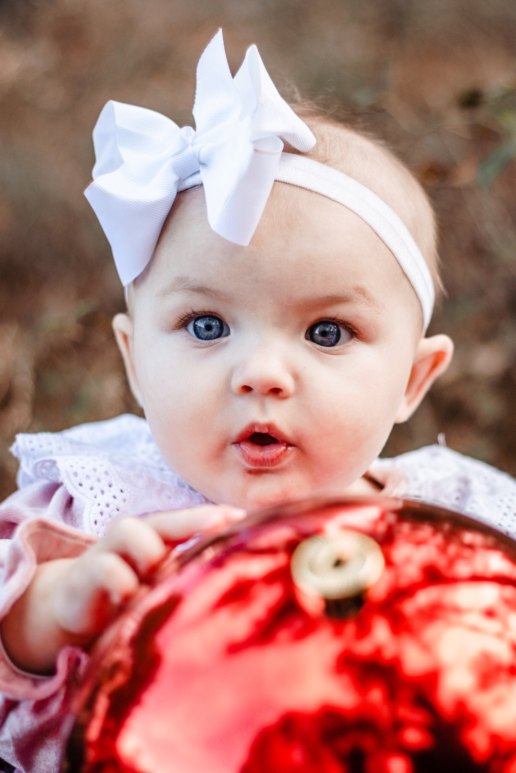 A baby with a white bow on her head is holding a red christmas ornament.