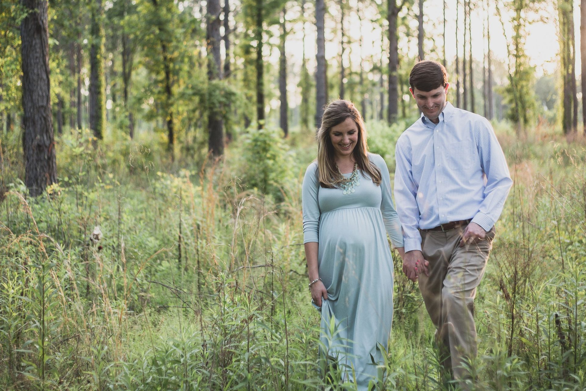 A man and a pregnant woman are walking through a forest holding hands.