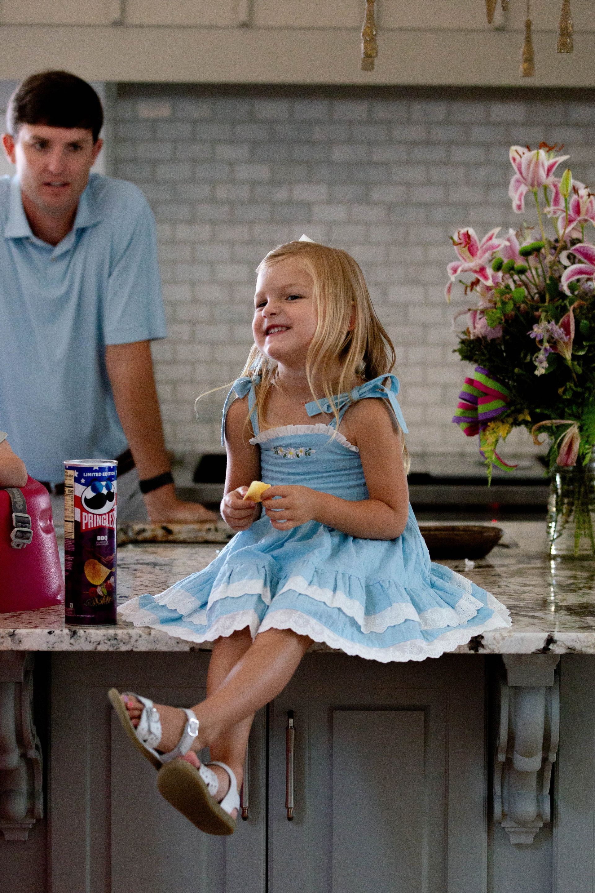 A little girl in a blue dress is sitting on a kitchen counter.